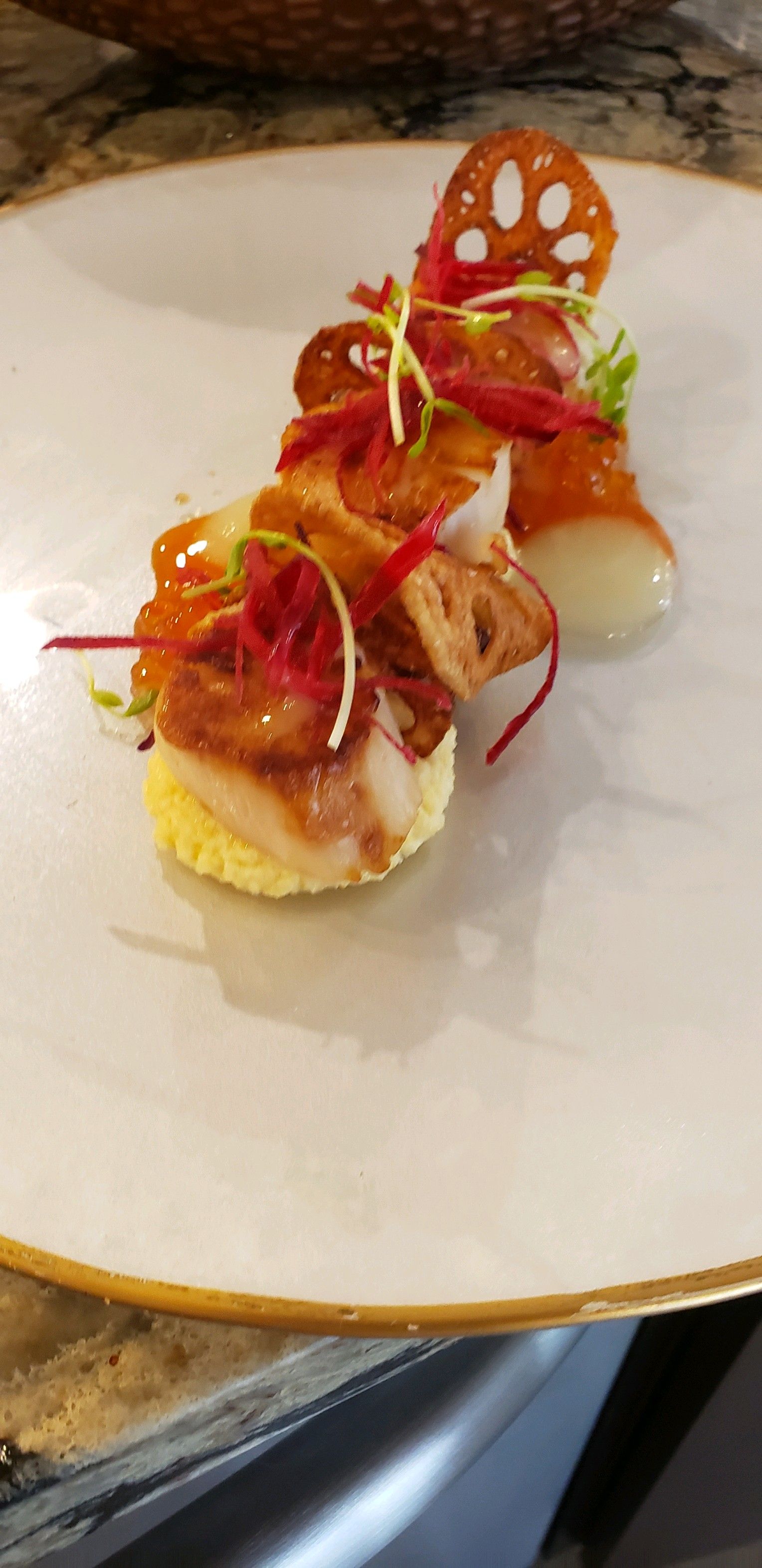 Gourmet dish with seared scallops on a yellow cake, topped with vegetables and lotus root chips.