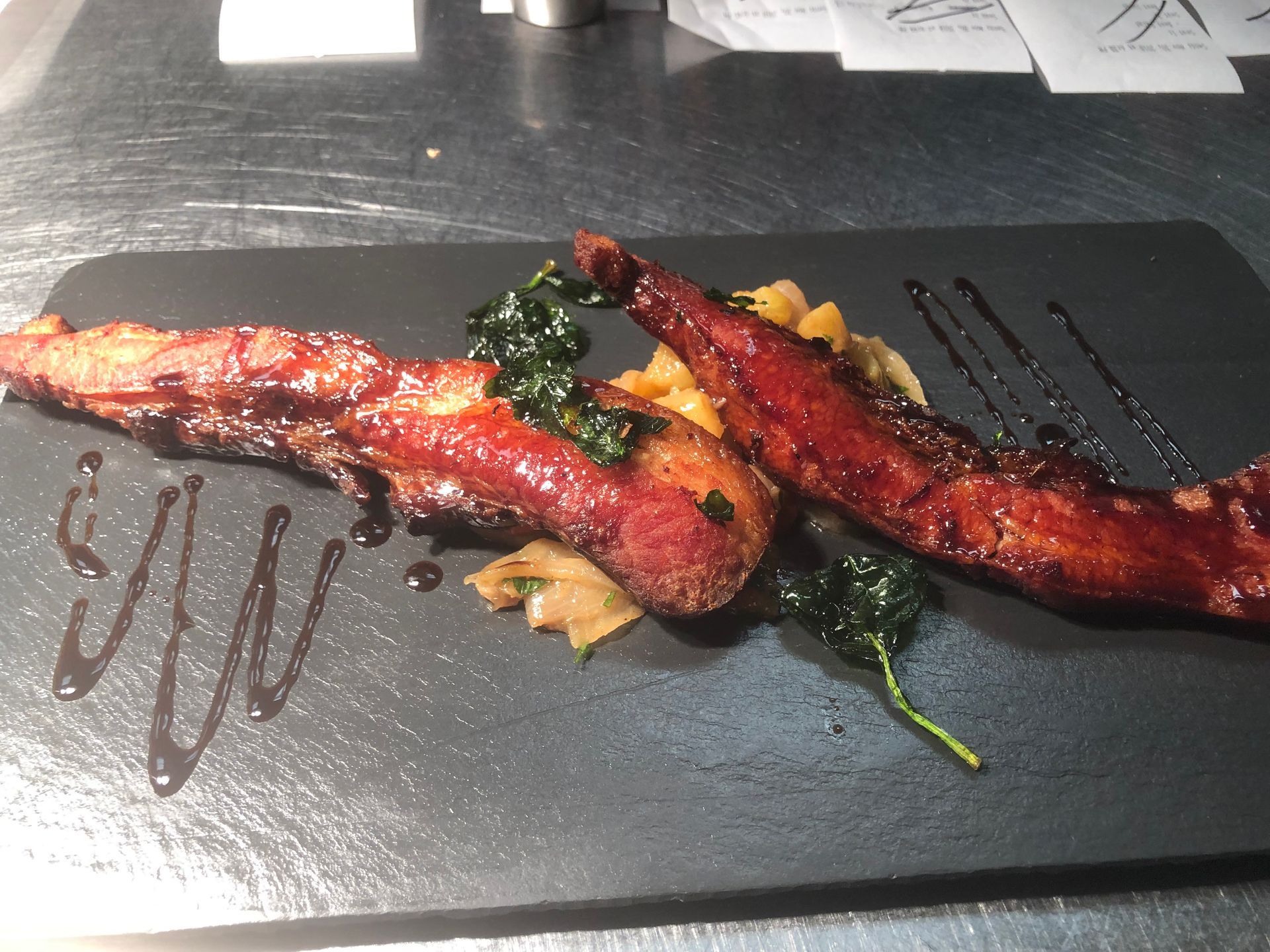 Two strips of glazed meat with sauce and greens on a black rectangular plate.