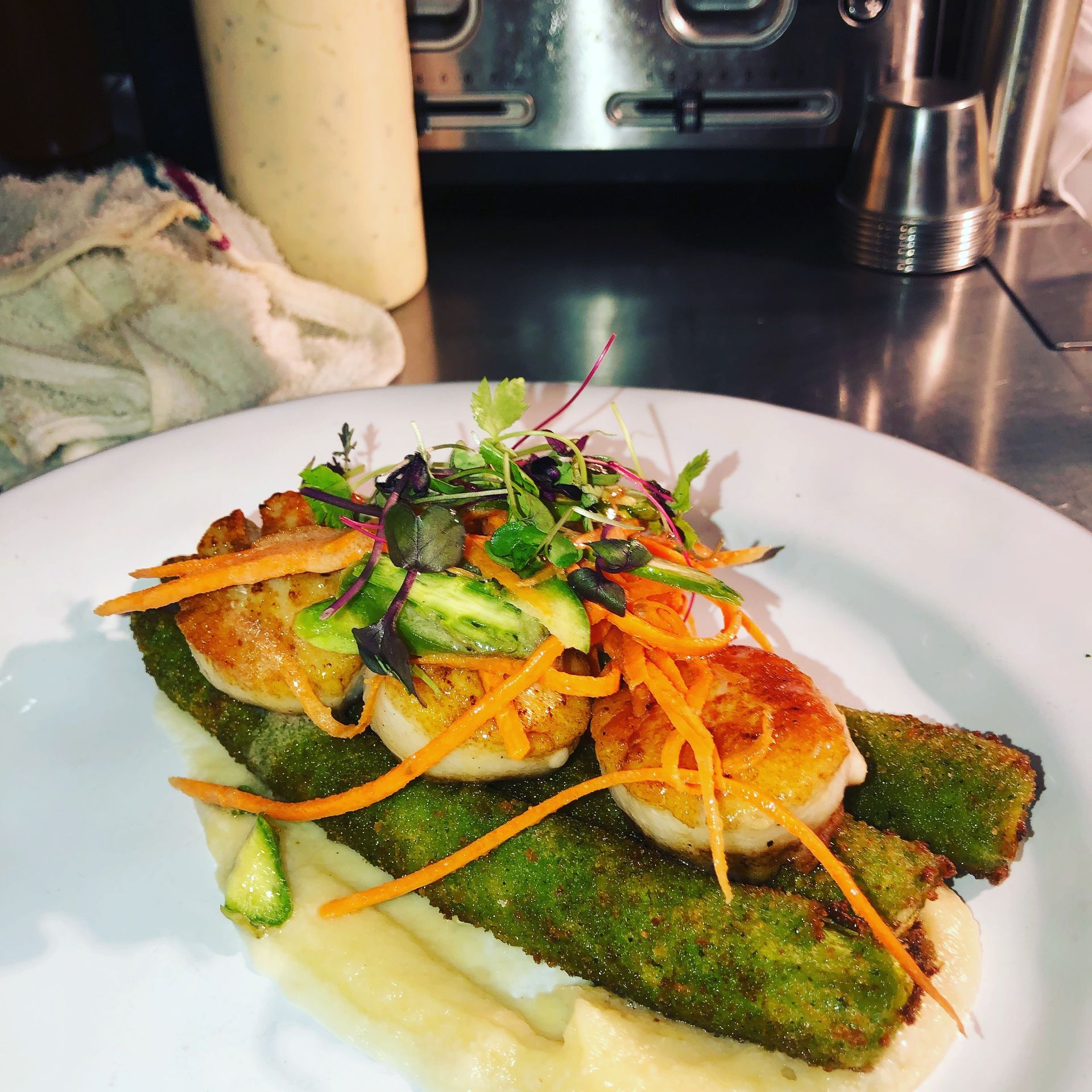 Seared scallops with green zucchini, carrots, avocado, and microgreens on a white plate.