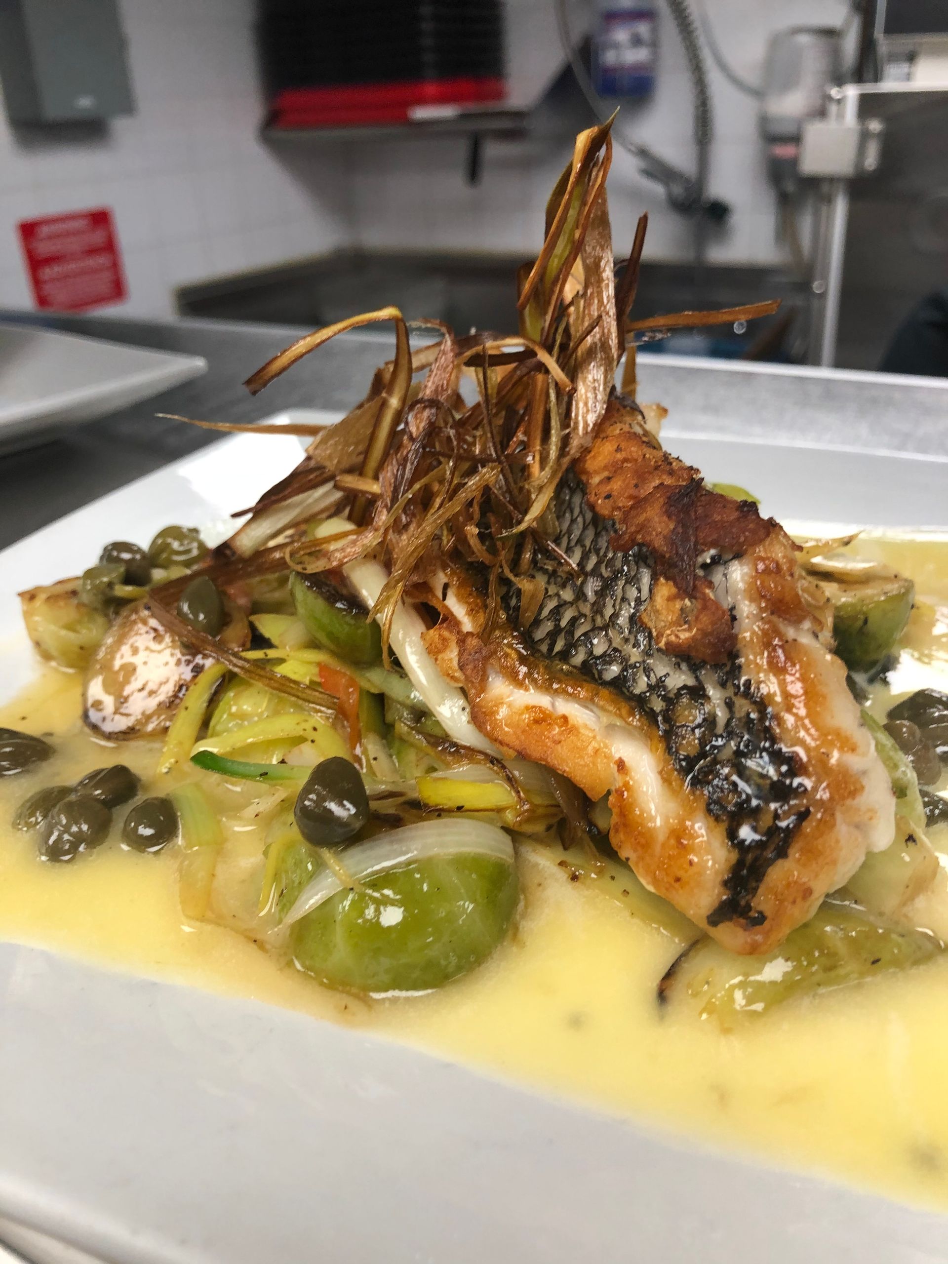 Pan-seared fish with fried fin, capers, and vegetables, in a light sauce on a white plate.