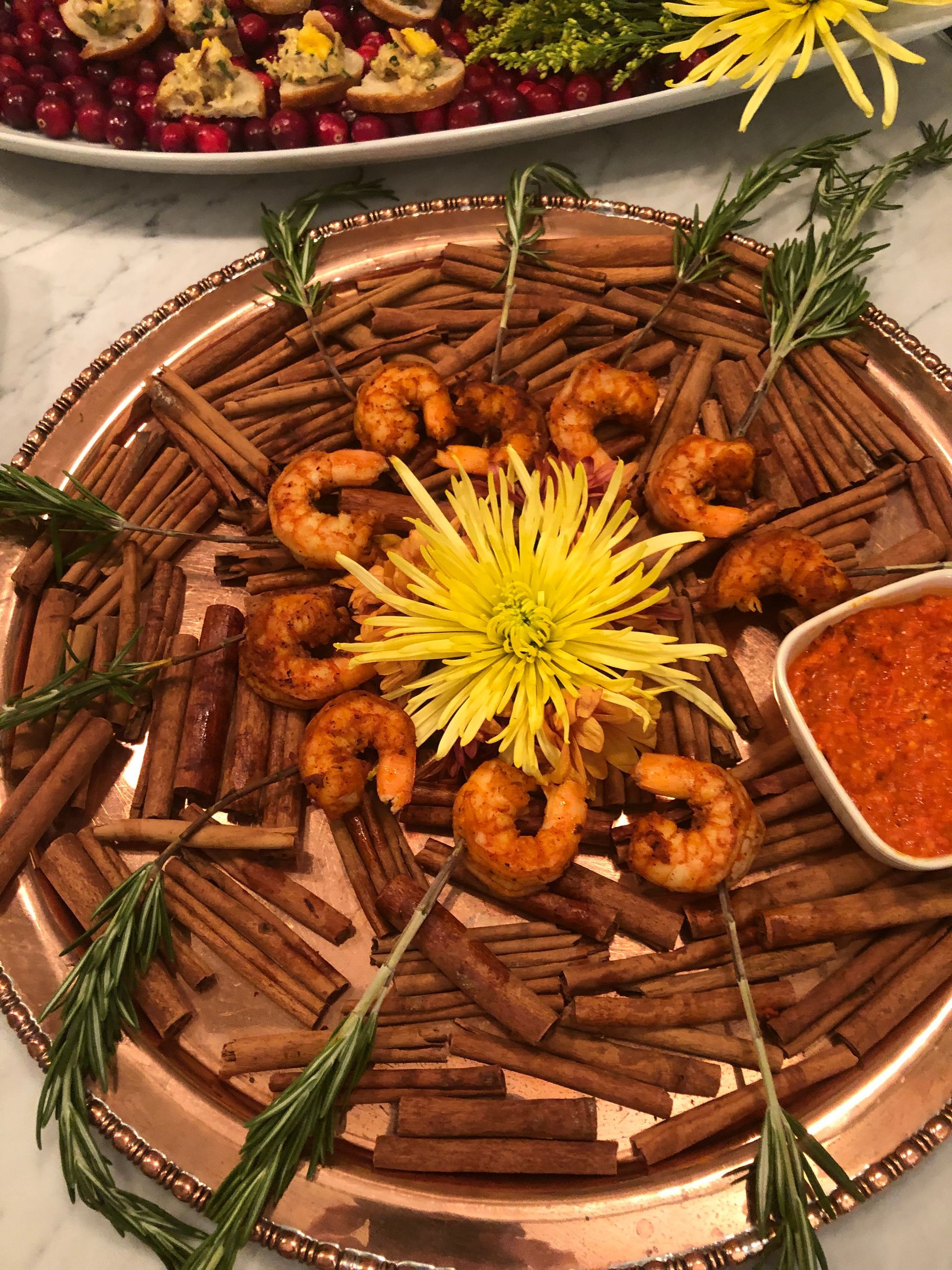 Shrimp appetizer platter with cinnamon sticks, rosemary, yellow flower, and dipping sauce.