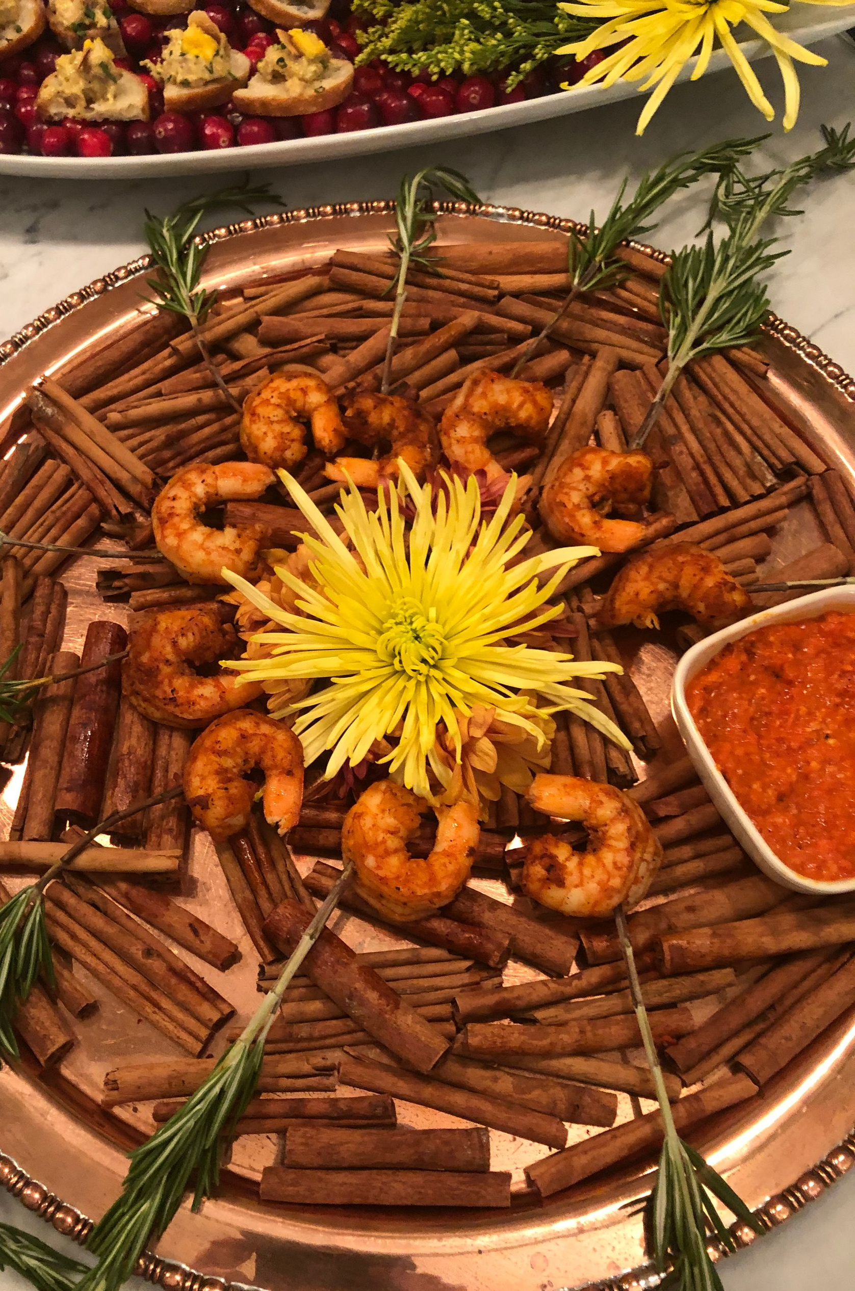 Spiced shrimp appetizer on a tray with cinnamon sticks and a flower, served with red sauce.