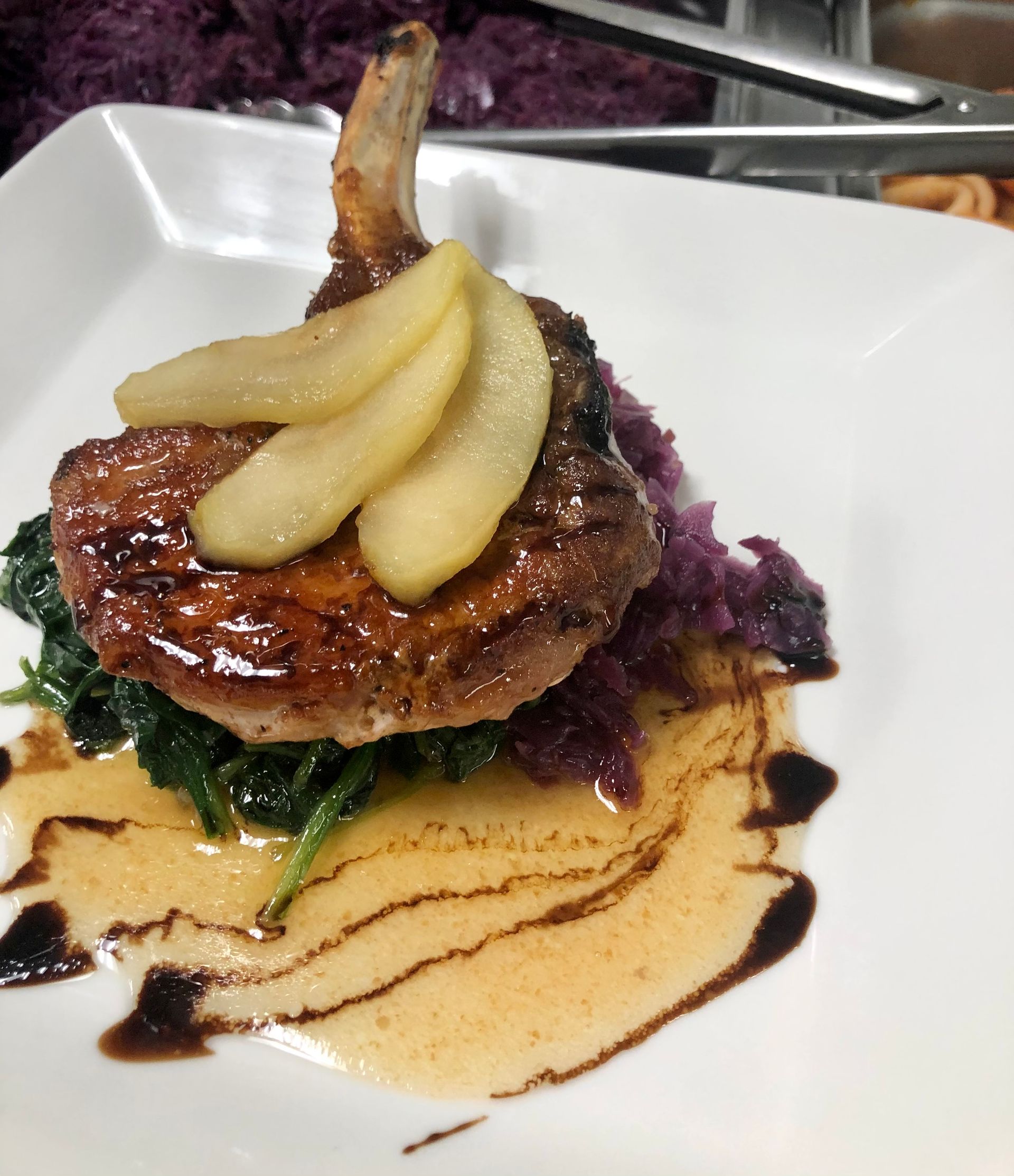 Seared pork chop with sauteed greens, pear slices, purple cabbage, and sauce, on a white plate.