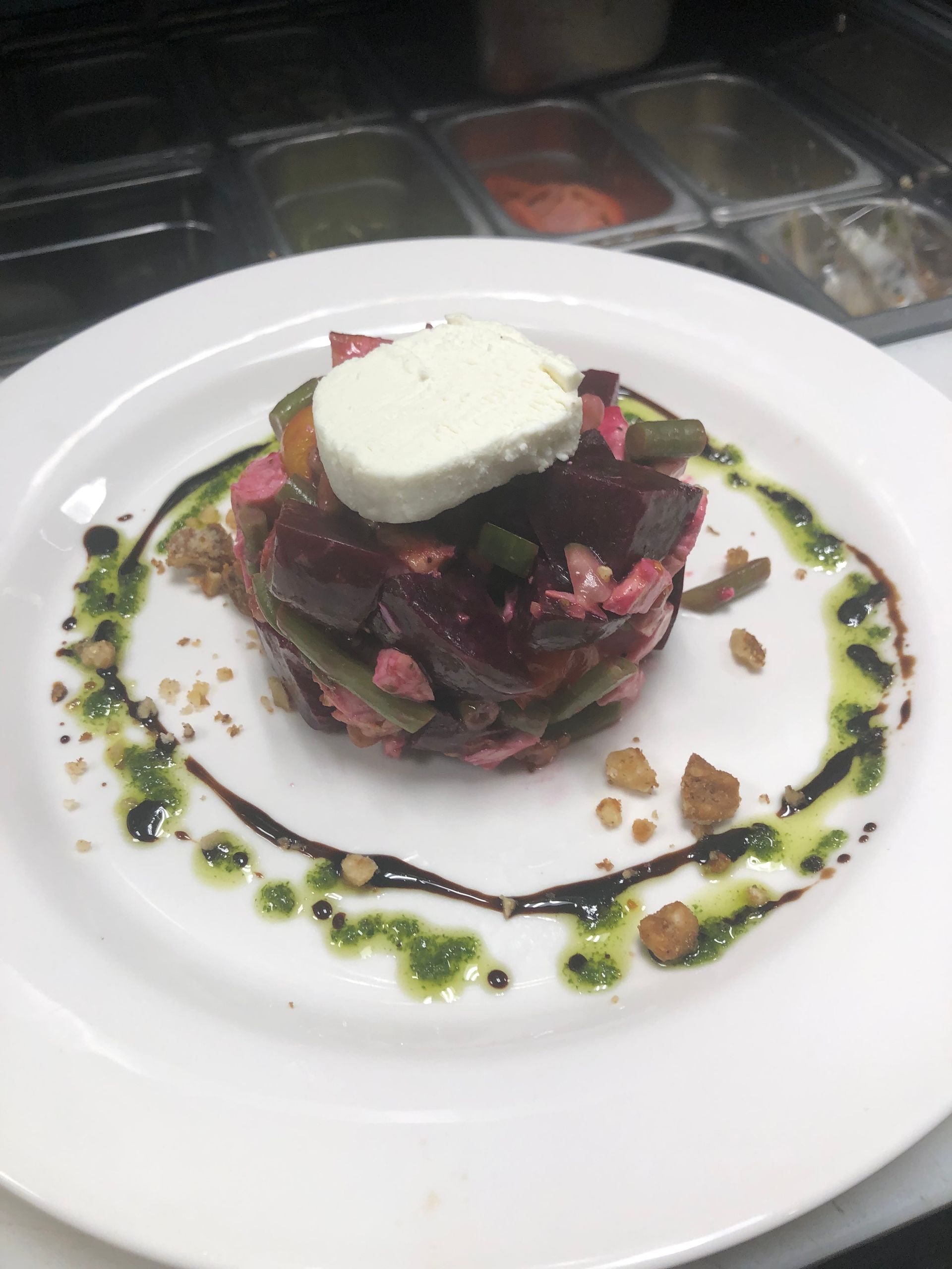 Stack of beet salad topped with cheese, drizzled with green sauce and balsamic glaze, on white plate.