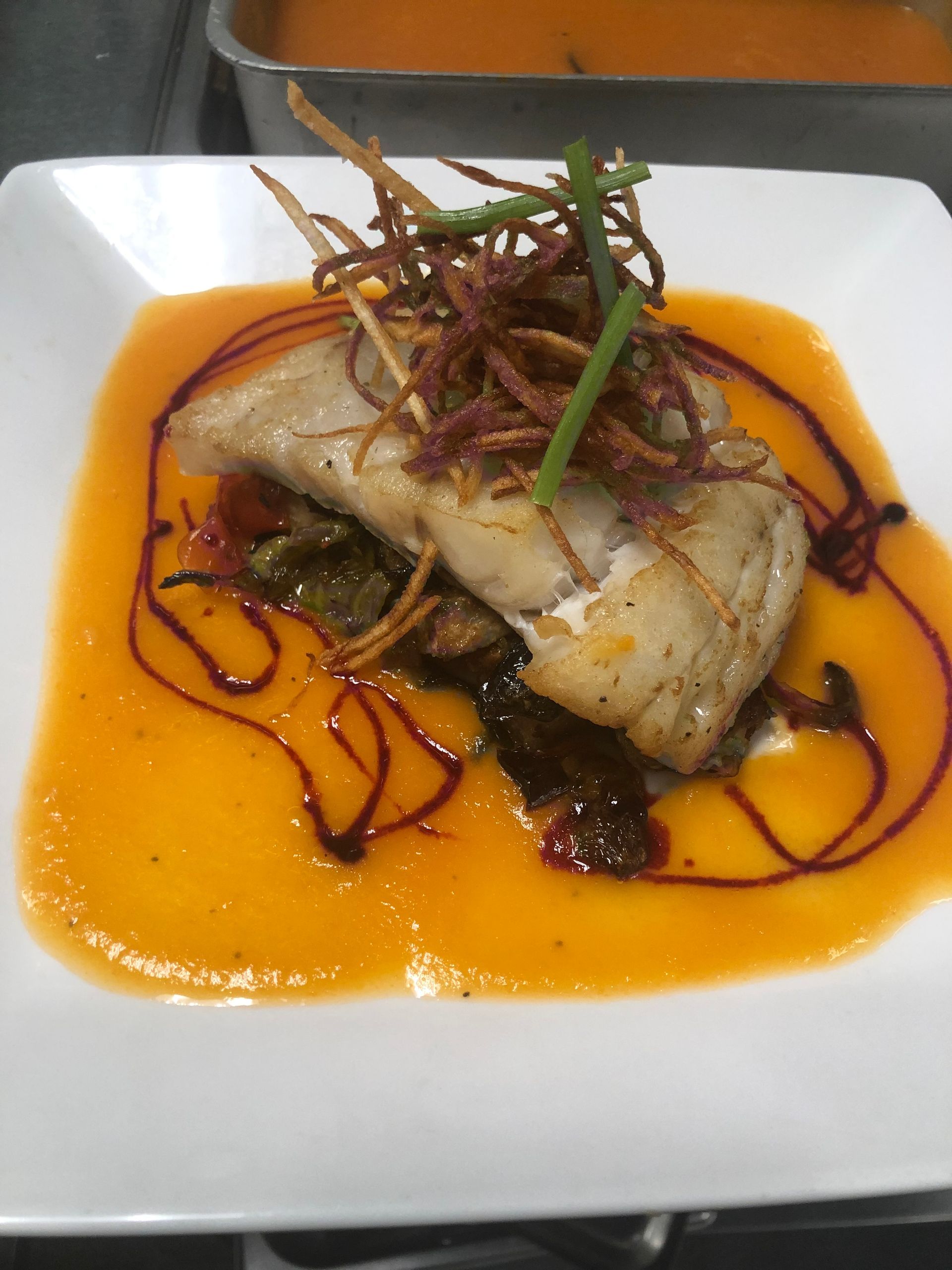 White plate with pan-seared fish on orange sauce, garnished with fried vegetables and chives.