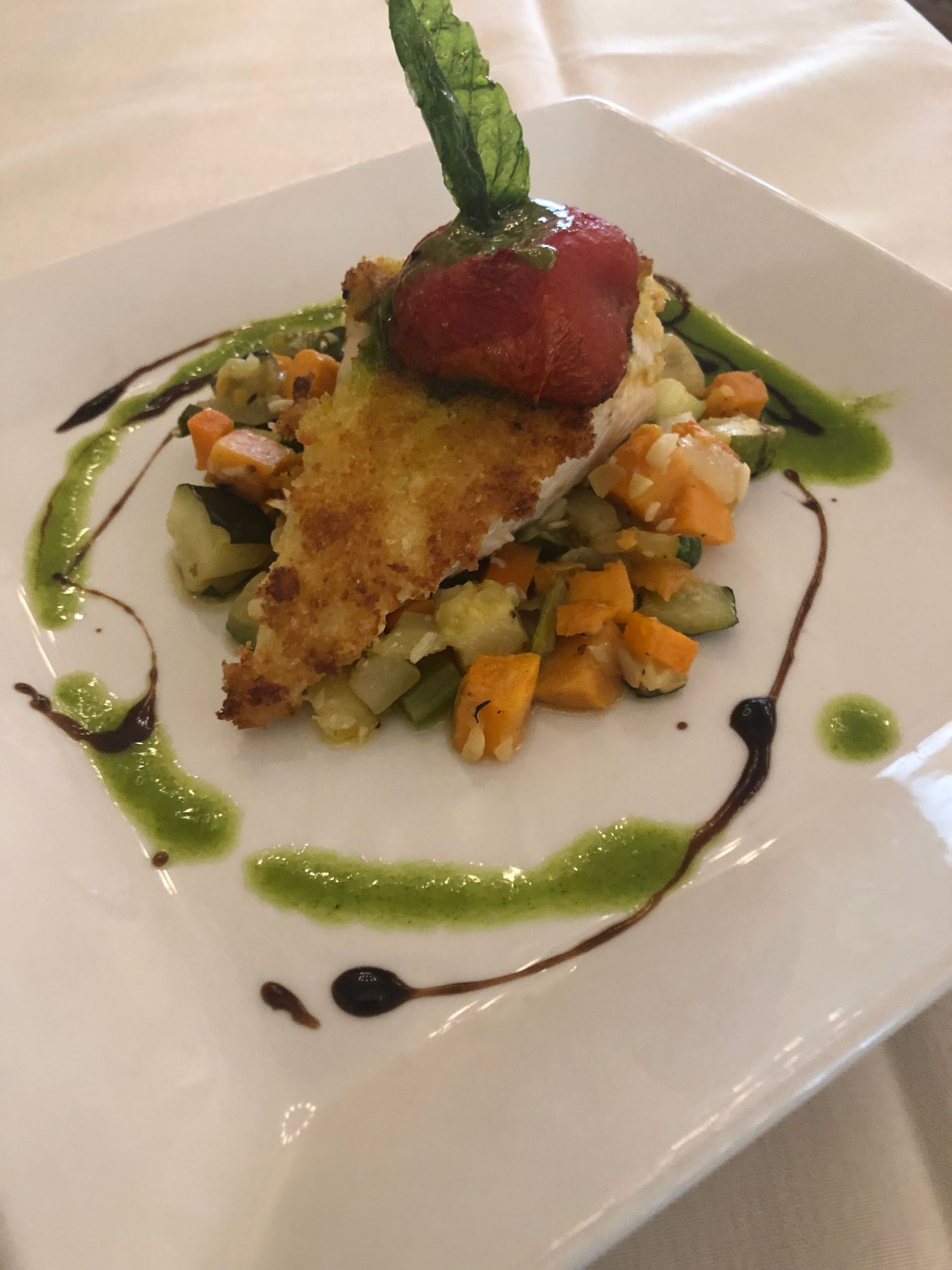 Breaded fish on vegetable medley, drizzled with green and black sauces. Garnished with mint and a red topping.