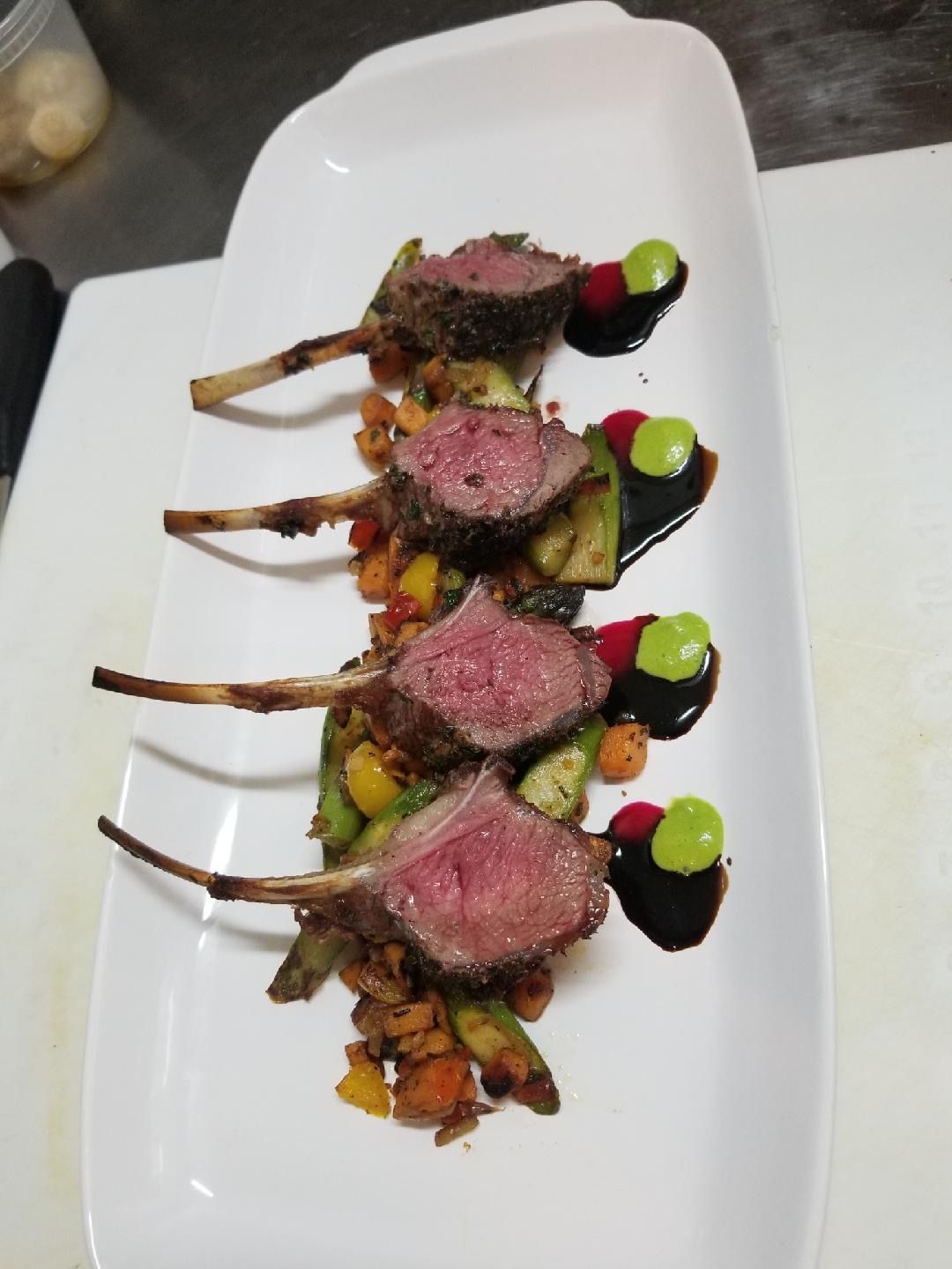 Rack of lamb on a bed of vegetables, drizzled with sauce and dots of green puree, served on a white platter.