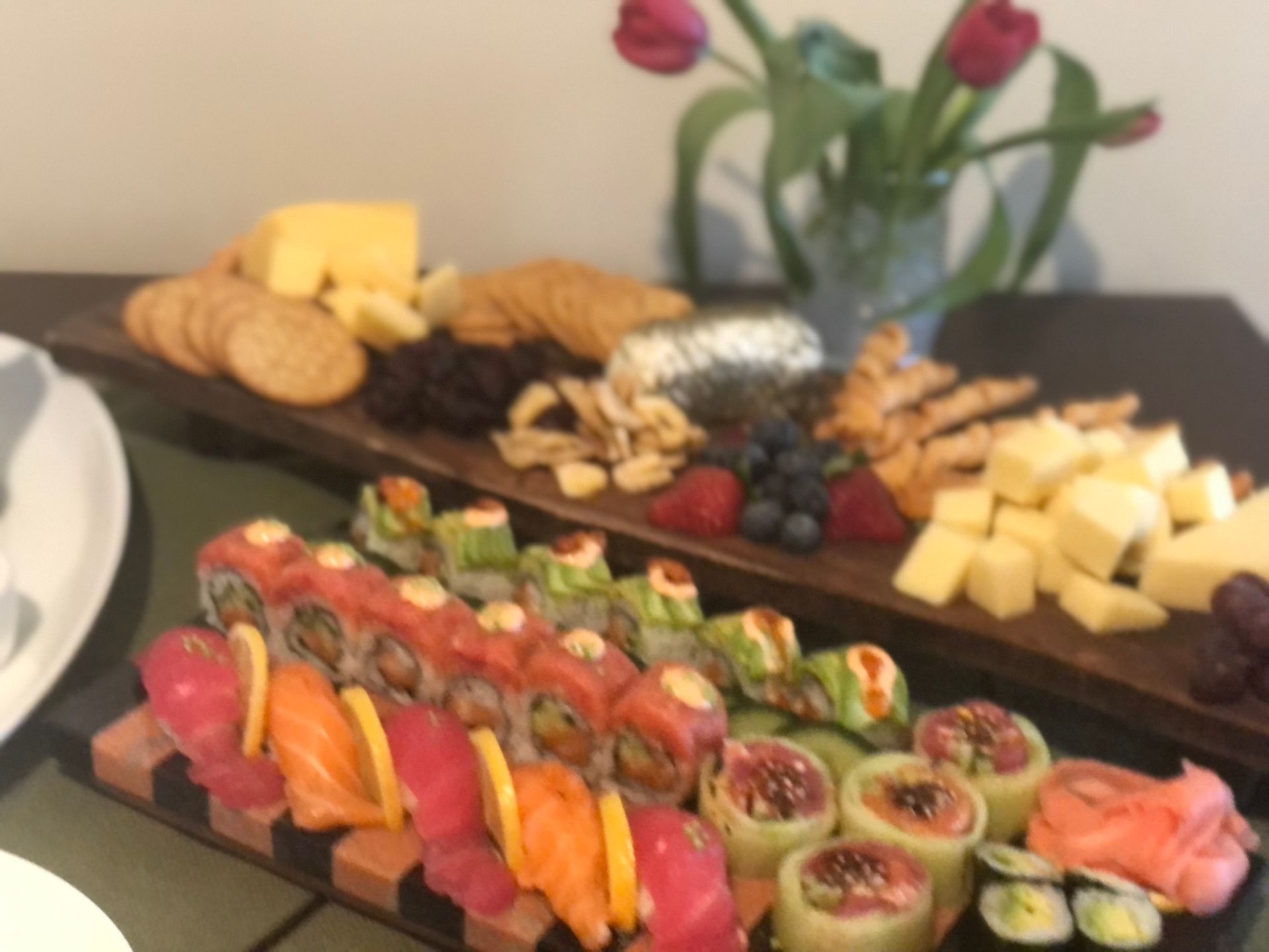 Sushi, cheese, and crackers on a wooden serving board with flowers in the background.
