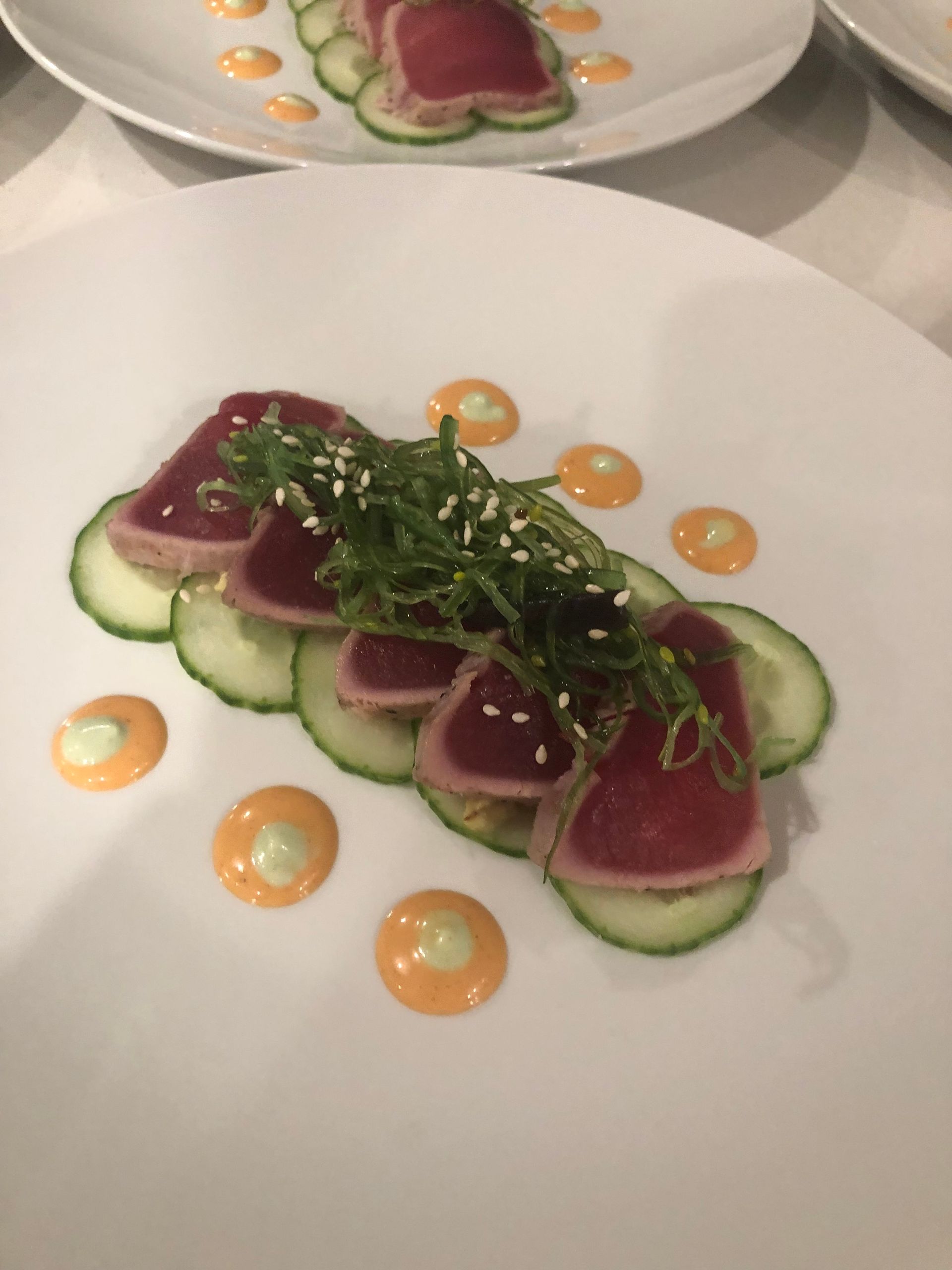 Seared tuna slices on cucumber, garnished with seaweed and sesame seeds, drizzled with orange sauce on white plate.