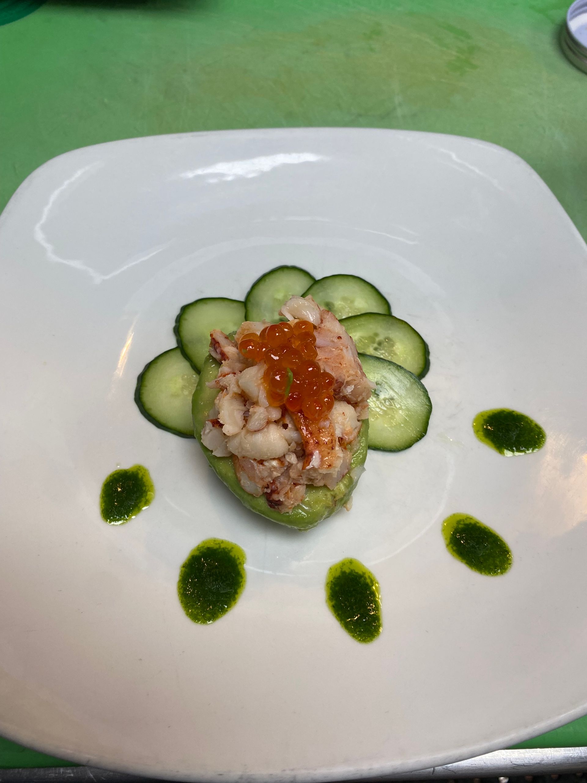 Lobster salad served in an avocado half, garnished with cucumber slices, salmon roe, and green sauce dots on a white plate.