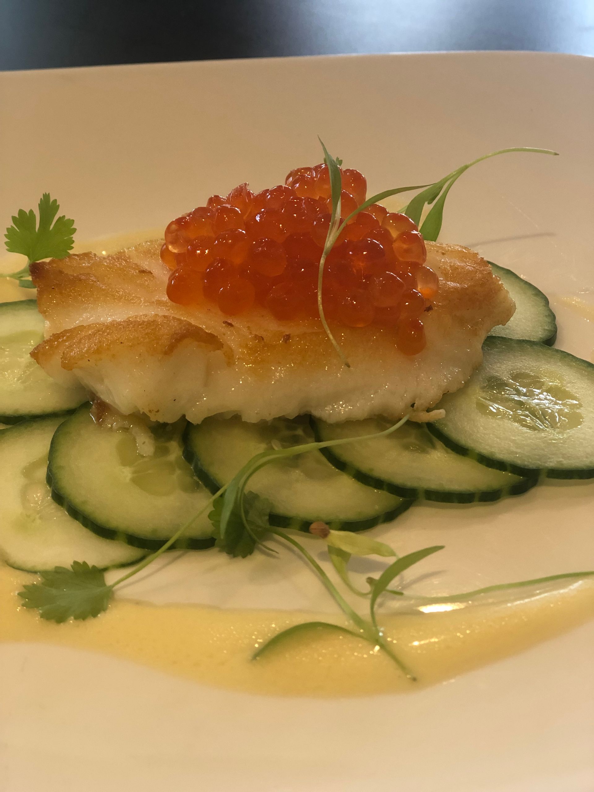 Pan-seared fish topped with red roe, cucumber slices, and herb sprigs on a white plate.