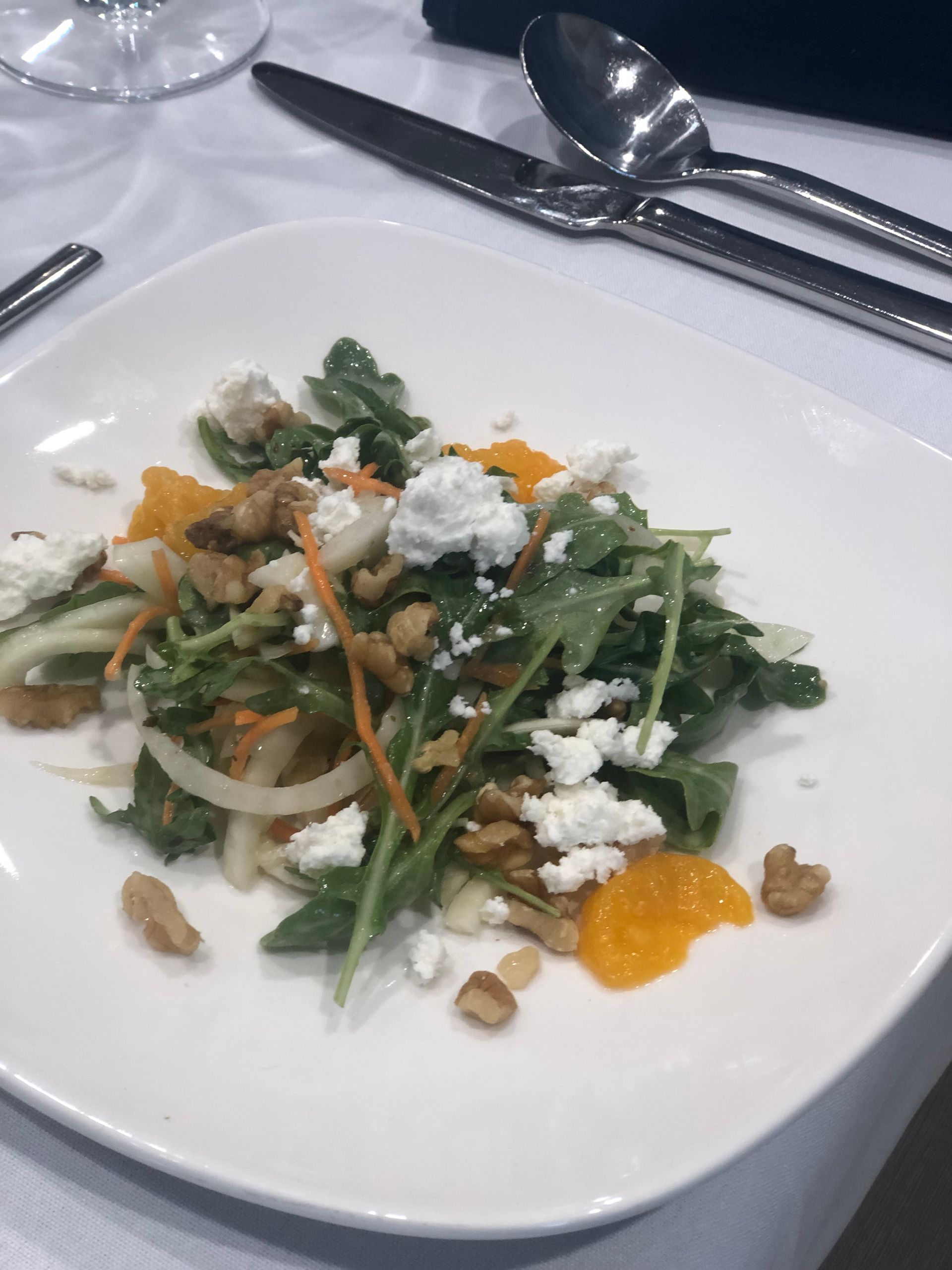 Salad on white plate: arugula, orange segments, goat cheese, walnuts; table setting with silverware.