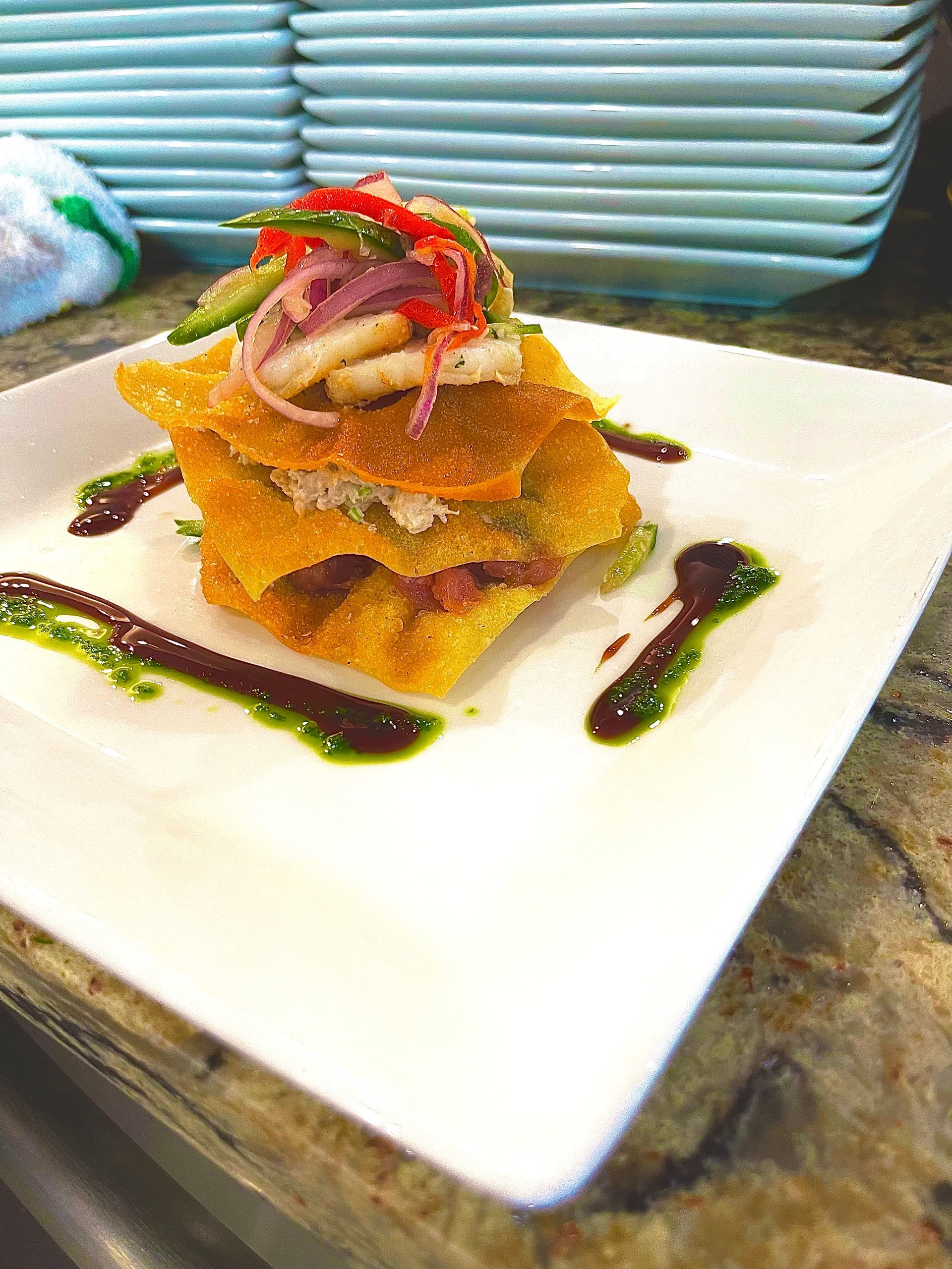 Stack of fried wontons layered with seafood and colorful vegetables on a plate, drizzled with green and brown sauces.