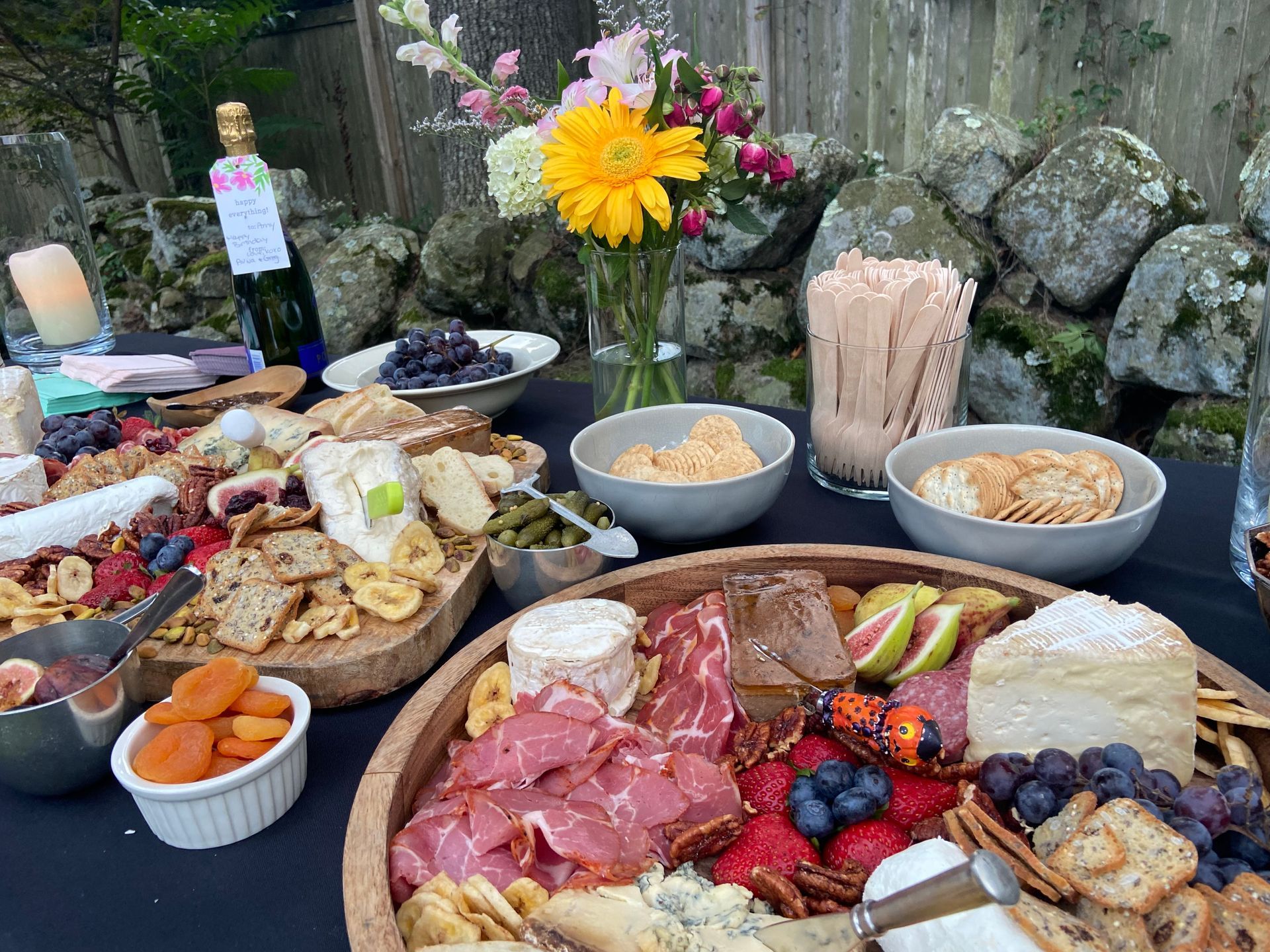 Charcuterie spread outdoors on a black table; cheeses, meats, fruit, crackers, flowers, and champagne.