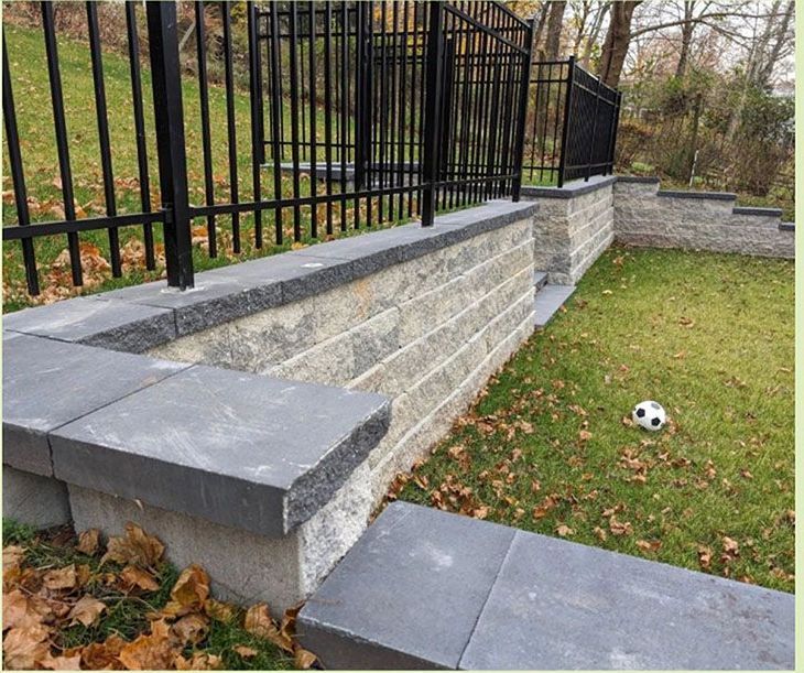 Retaining wall with black fence and gray stone, built on a slight slope; soccer ball on grass.