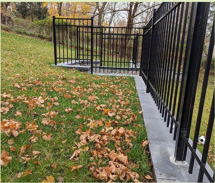 Black metal fence on a grassy slope with fallen leaves; a low retaining wall separates the grass.