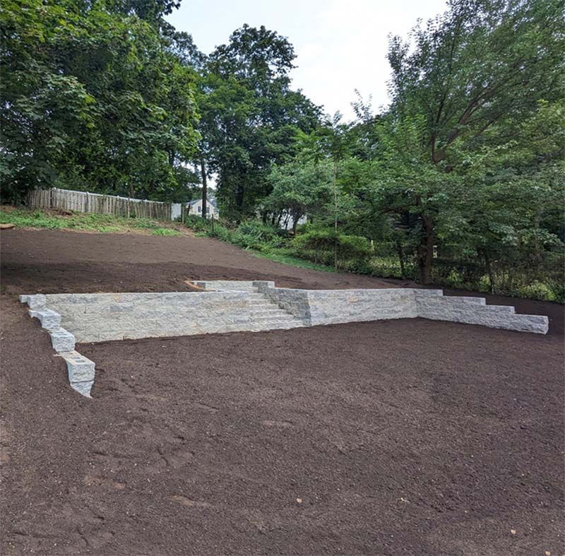 Stone retaining wall with steps on a sloped, freshly tilled yard, trees in background.