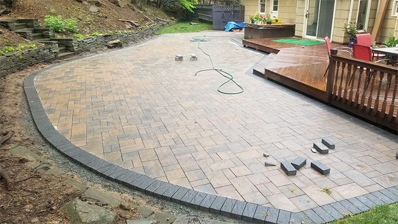 A newly constructed brick patio with a dark border, adjacent to a wooden deck and a house.