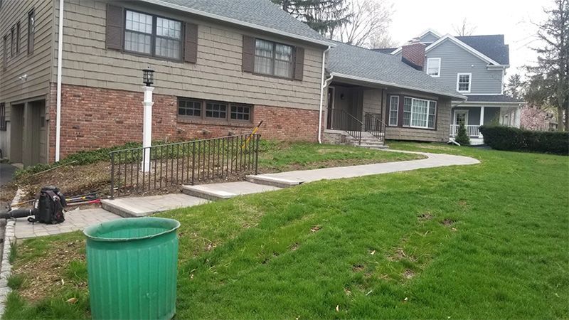 Green trash can, walkway, and houses with brown and gray siding, with grass.