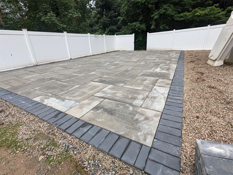 A paved patio enclosed by a white fence with a dark brick border, surrounded by gravel.