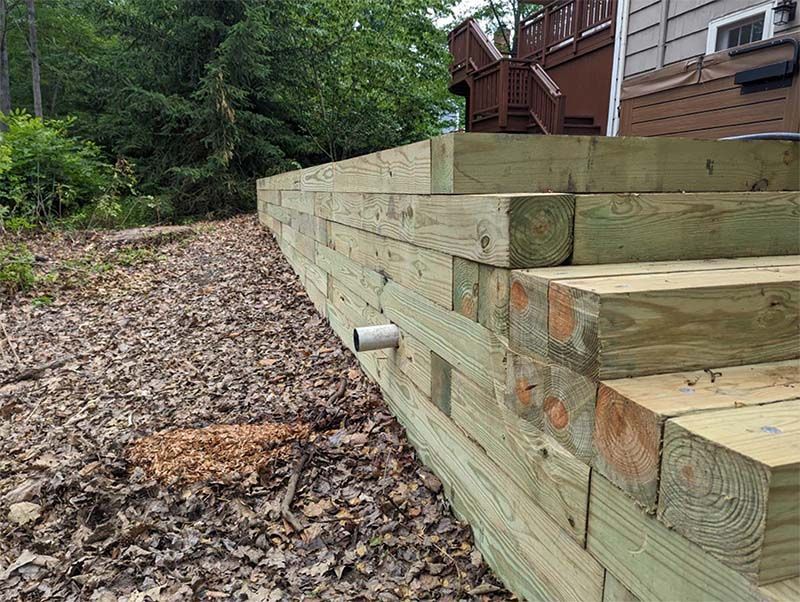 Wooden retaining wall and steps beside a wooded area, with a deck in the background.