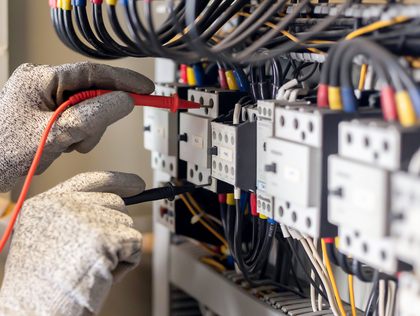 Electrician in gloves tests wiring with a multimeter inside an electrical panel.
