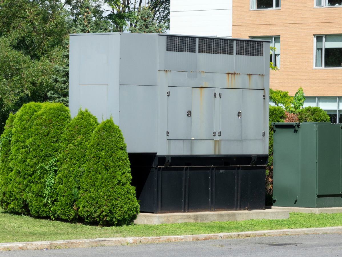 Large grey electrical box behind green bushes, next to a building.