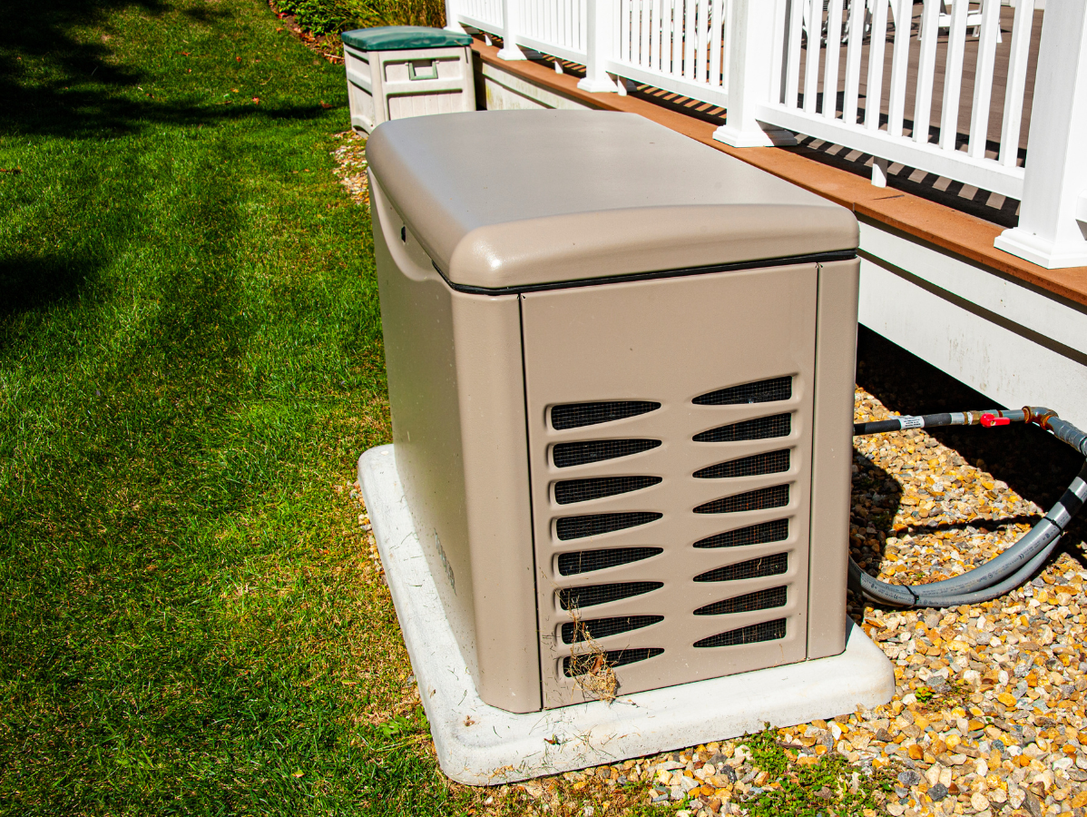 Tan generator on a concrete pad next to a house with white railings, on a lawn.