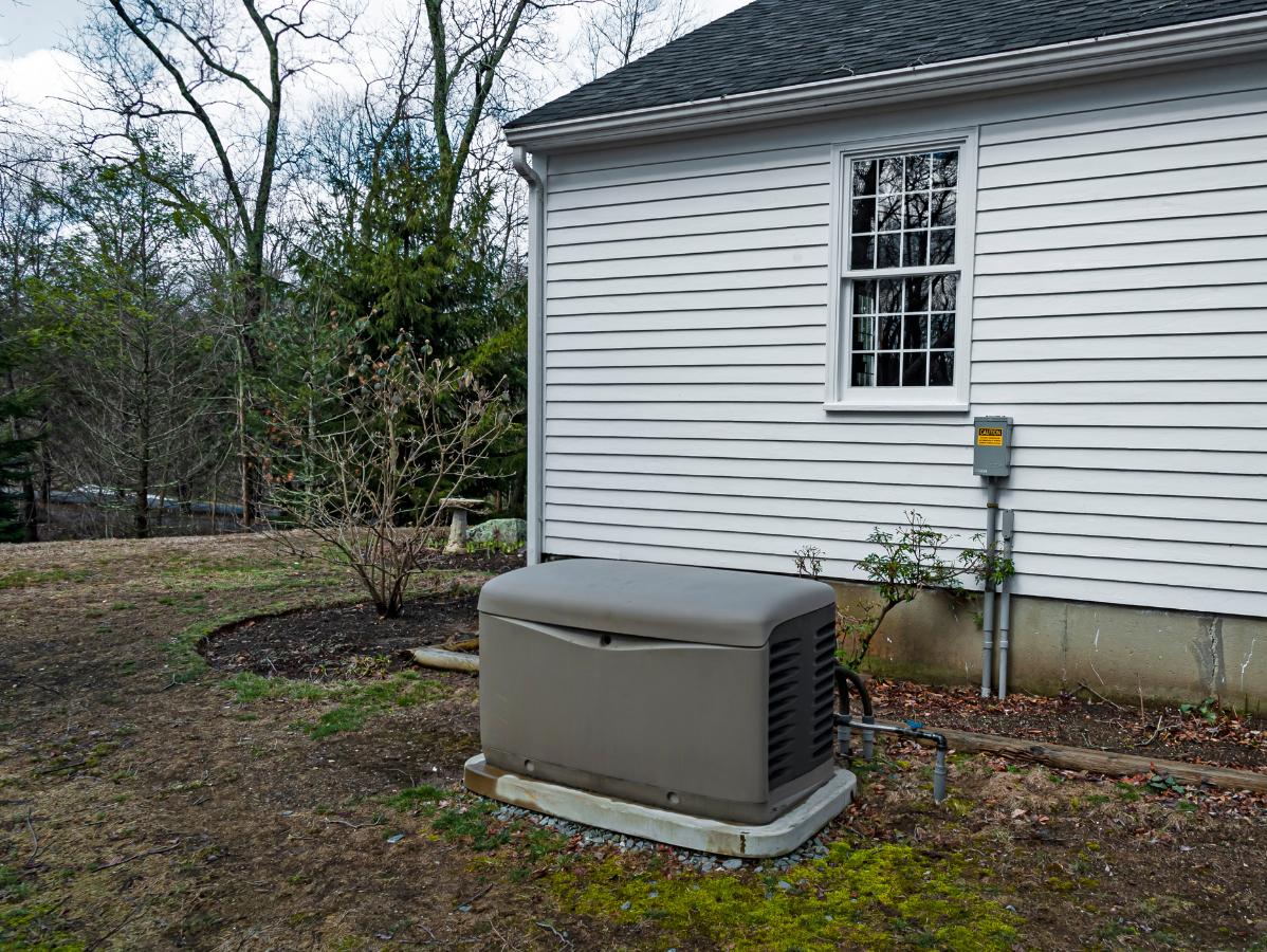 Generator near a white house, on a concrete pad. Electrical box on the wall. Brown, green surroundings.