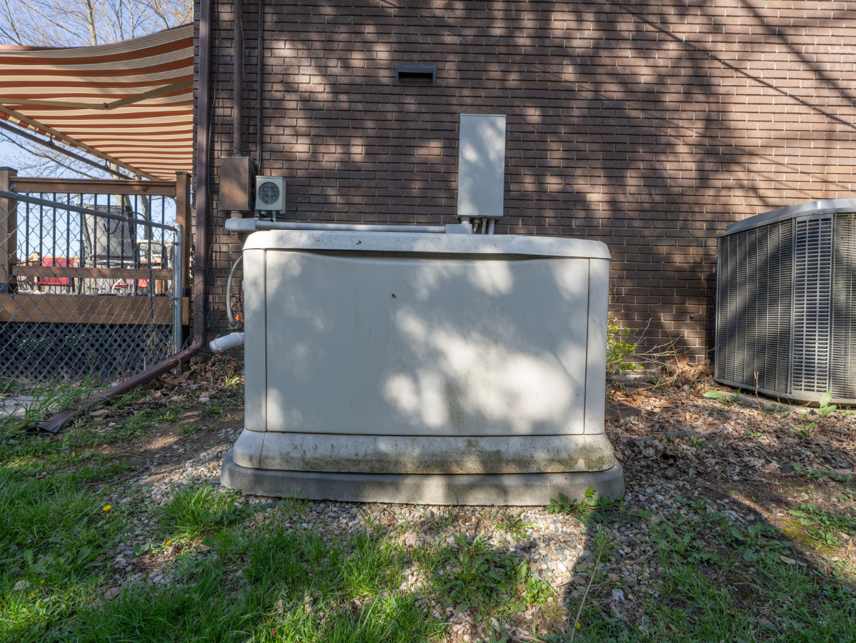 Large, beige generator on concrete base against a brick wall, with air conditioning unit to the right.