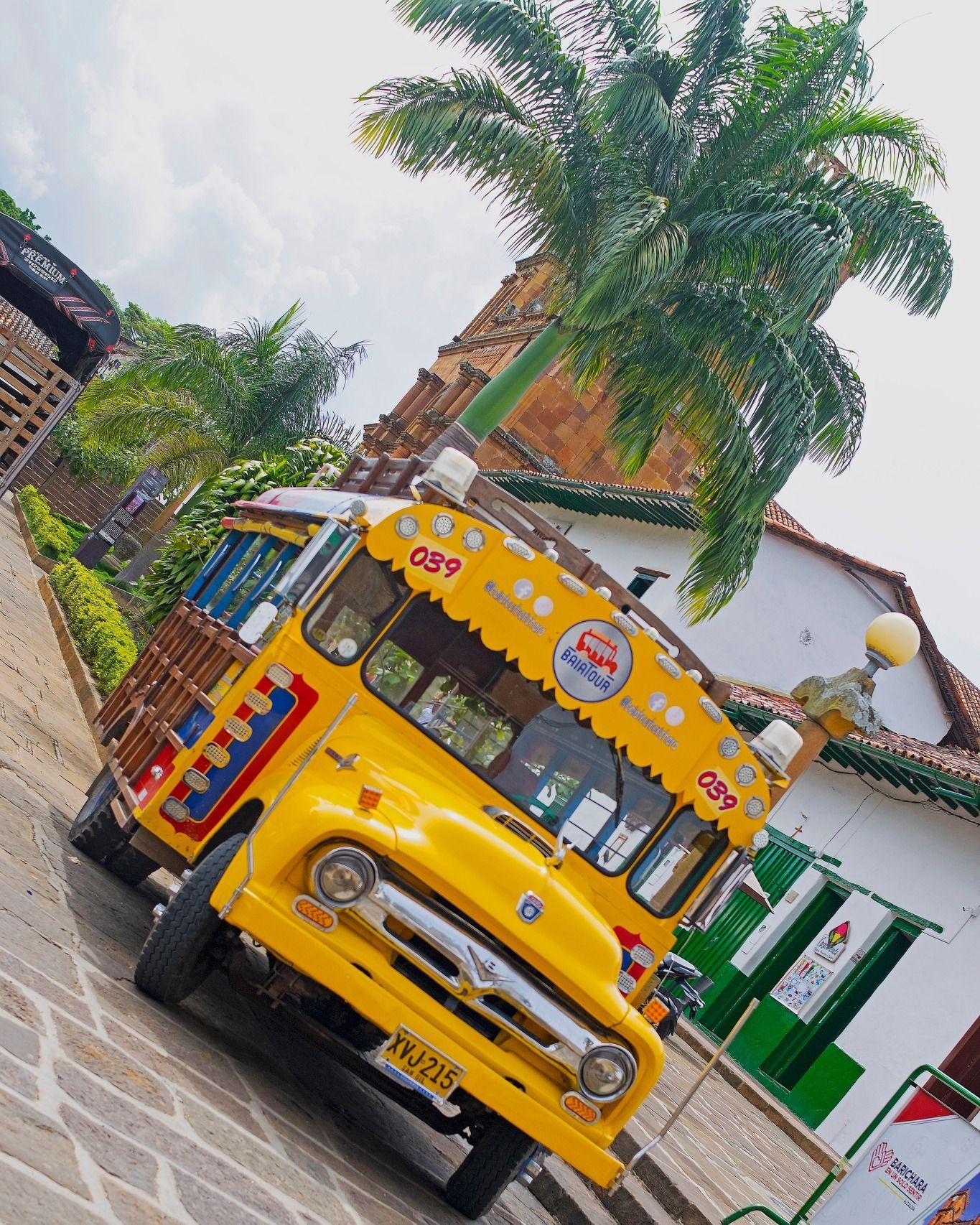 Autobús amarillo decorado con vibrantes colores estacionado en una calle adoquinada. Palmera y edificio al fondo.