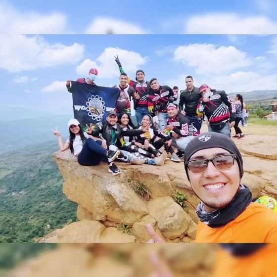 Grupo de personas en un acantilado, con una bandera, sonriendo. Montaña y cielo al fondo.