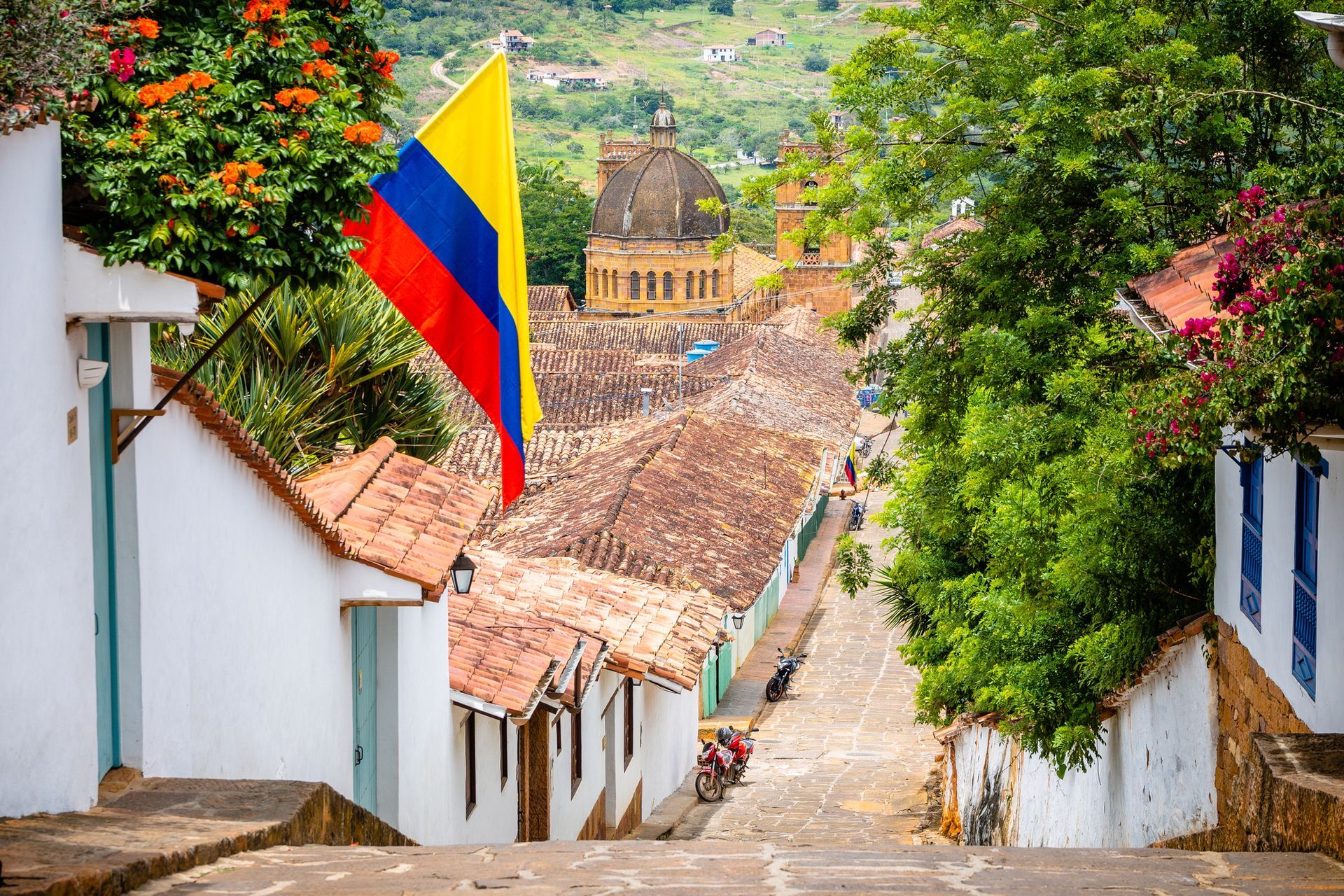 Bandera colombiana ondeando sobre una calle adoquinada bordeada de edificios blancos, techos de tejas y vegetación.