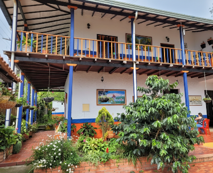 Edificio blanco de dos pisos con detalles en azul y naranja, balcones y exuberante vegetación.