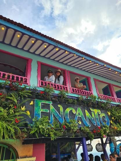 Edificio blanco de dos pisos con detalles en azul y naranja, balcones y exuberante vegetación.