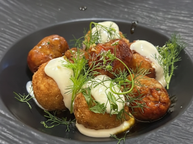 Comida frita en un recipiente oscuro, rociada con salsa blanca y guarnición de eneldo.