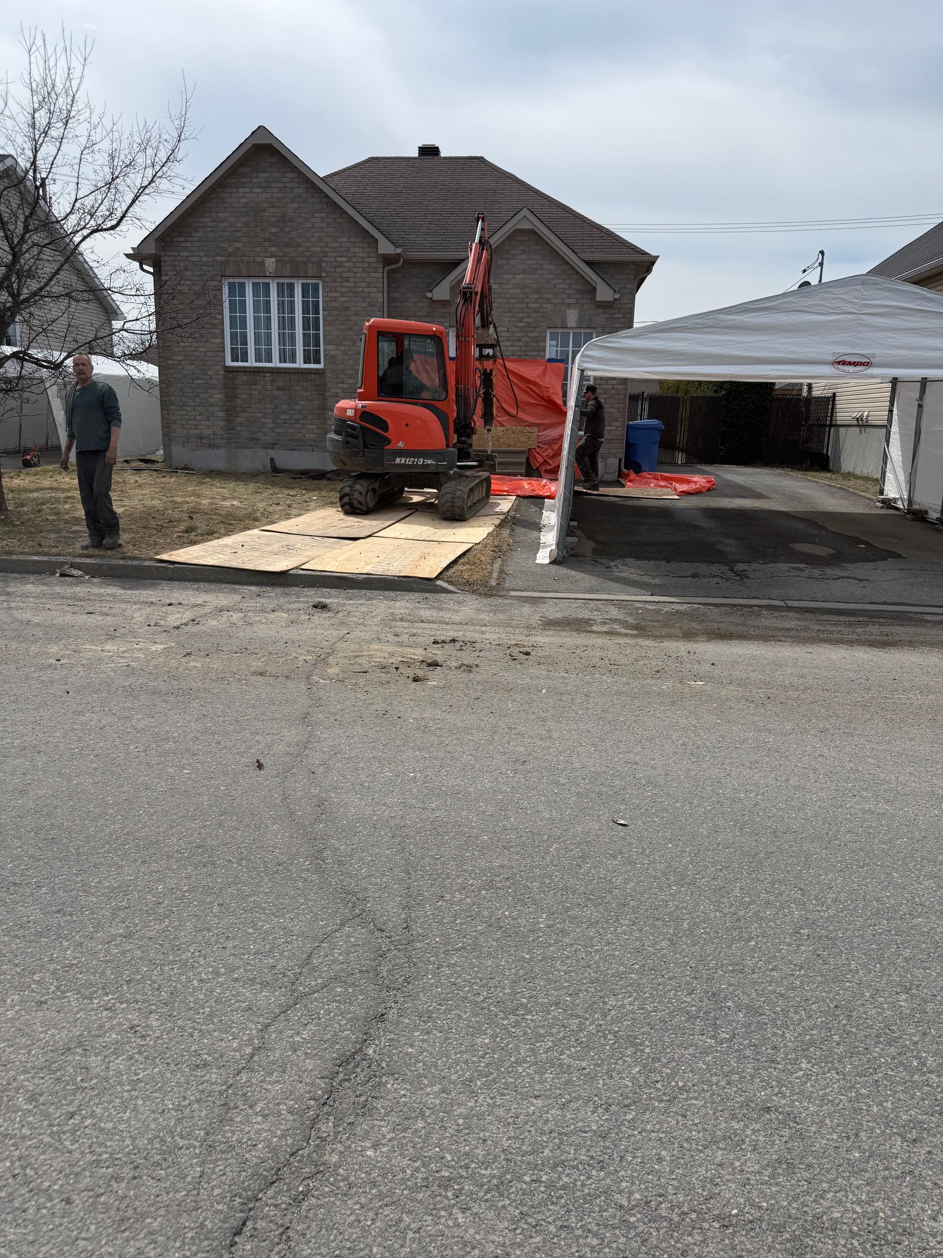 Un homme se tient devant une maison avec une excavatrice garée devant.
