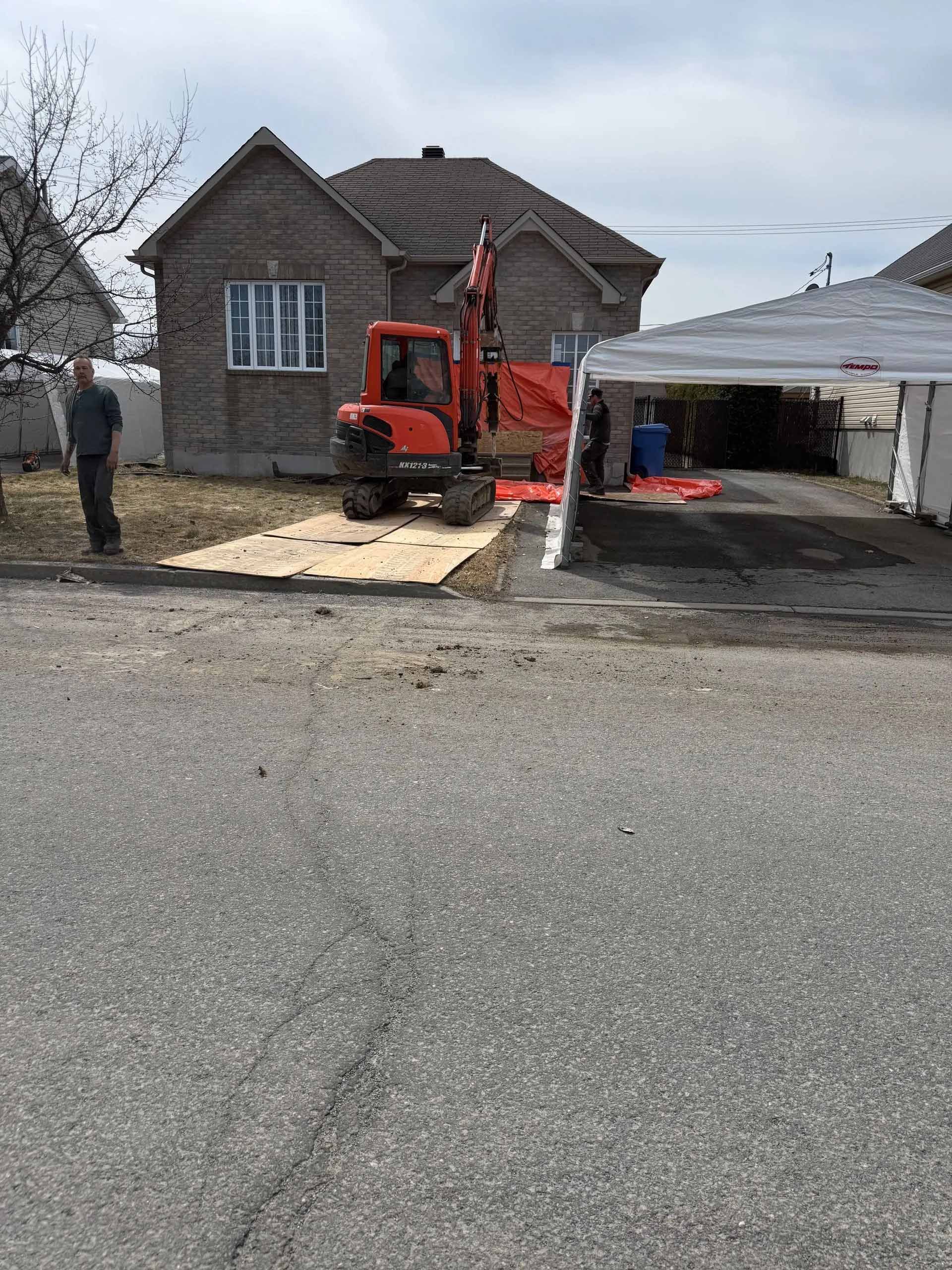 Un homme se tient devant une maison avec une excavatrice garée devant.