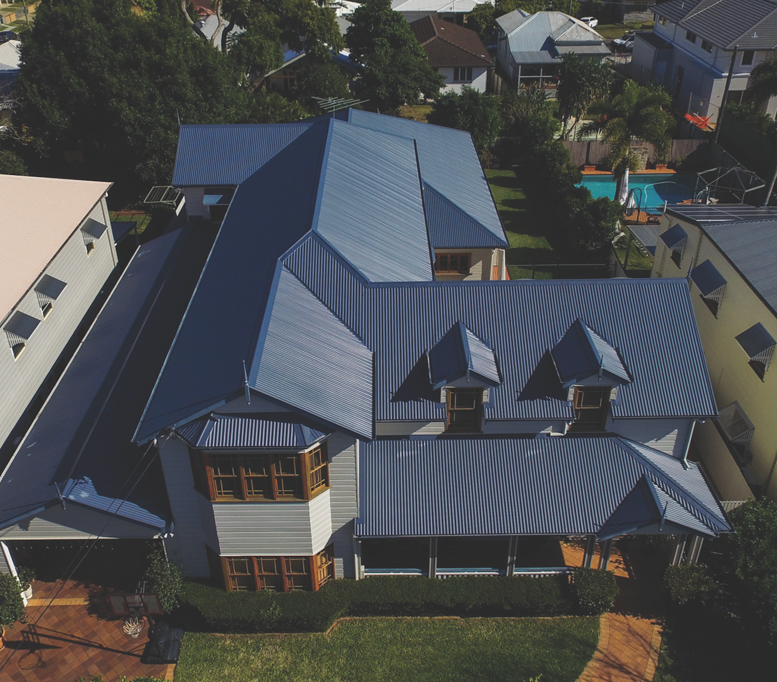 Aerial view of a large house with a dark blue metal roof and surrounding trees in a suburban neighborhood