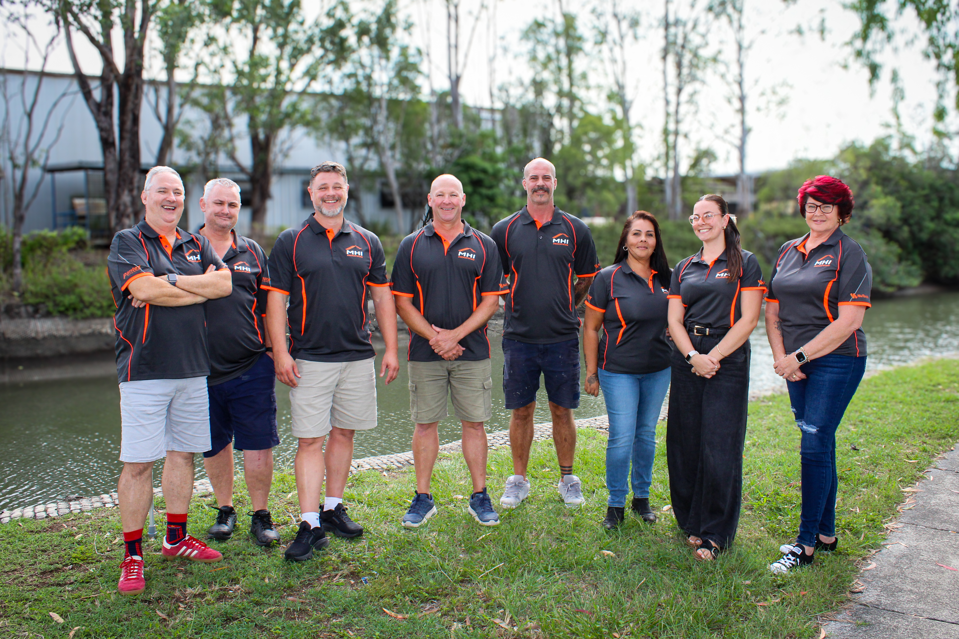 Seven people in black shirts standing by a pond on a grassy path, smiling outdoors