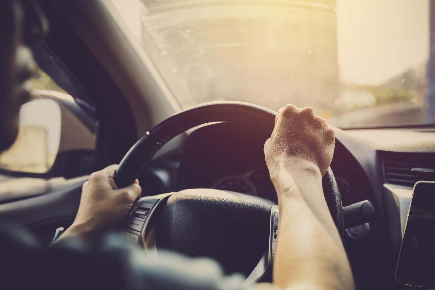 Hands gripping a steering wheel inside a car, driving in sunlight.