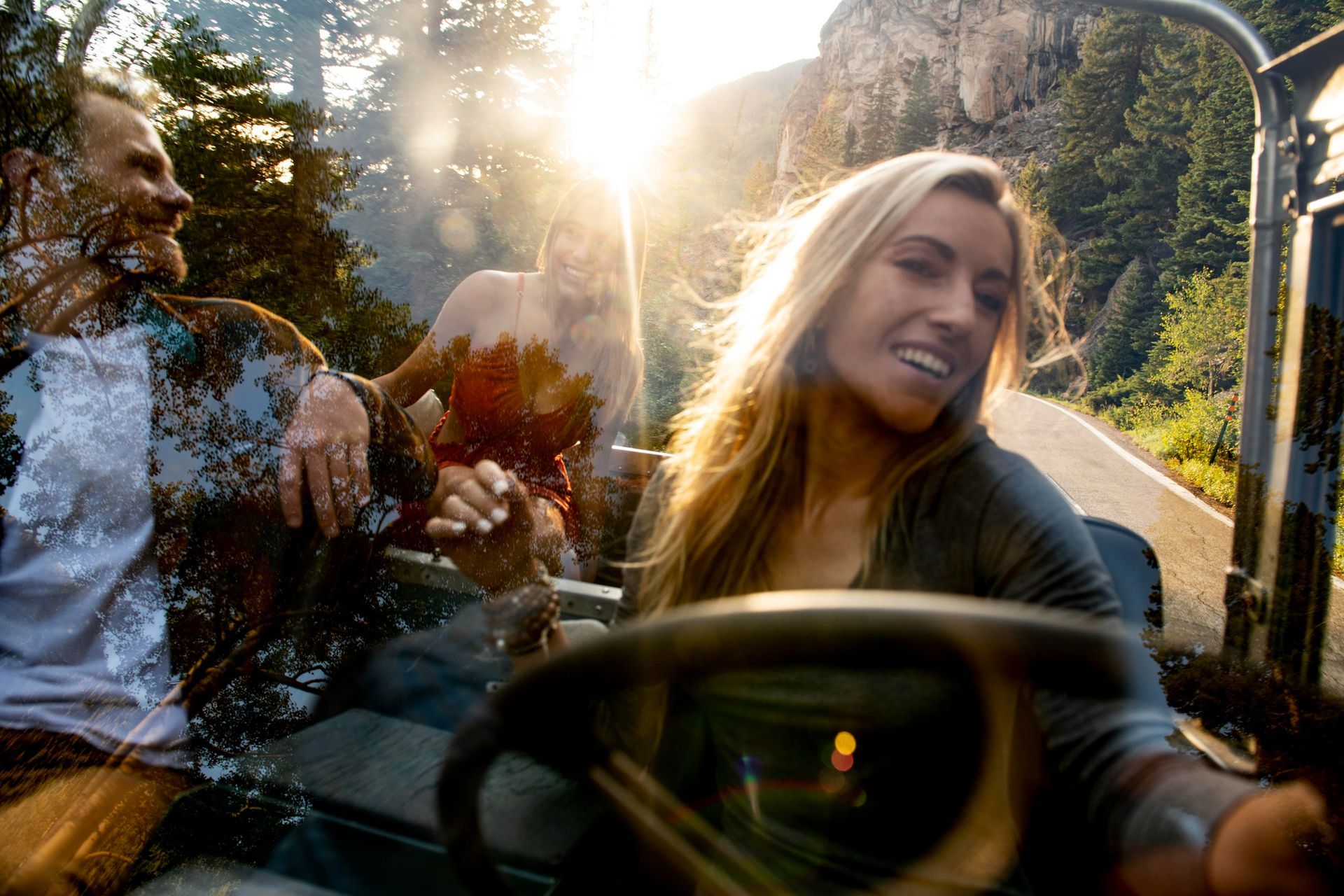 Three people in an open-top vehicle on a scenic road with sunlight. One is driving, smiling.