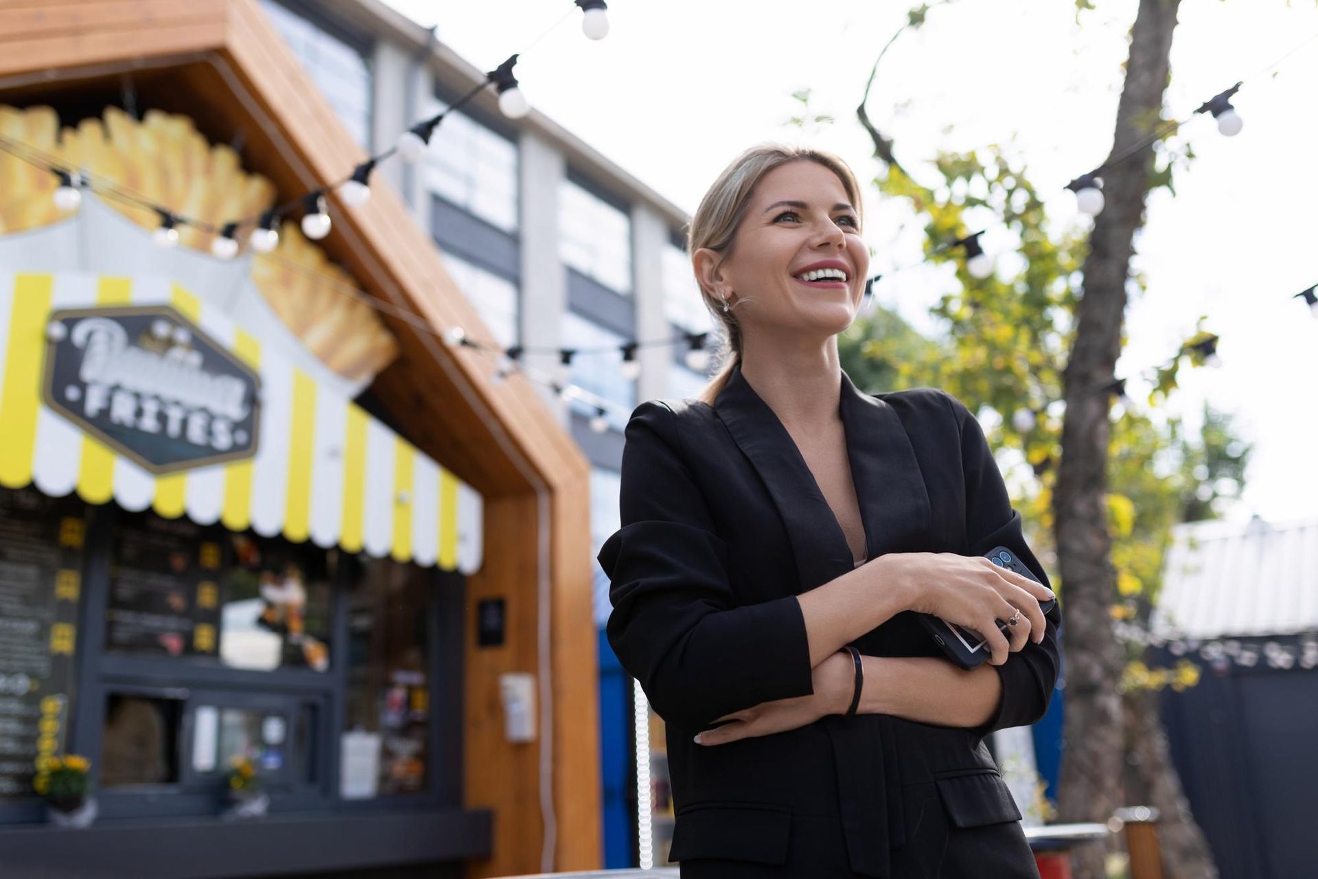 Woman in black blazer smiles, standing near a food stall.