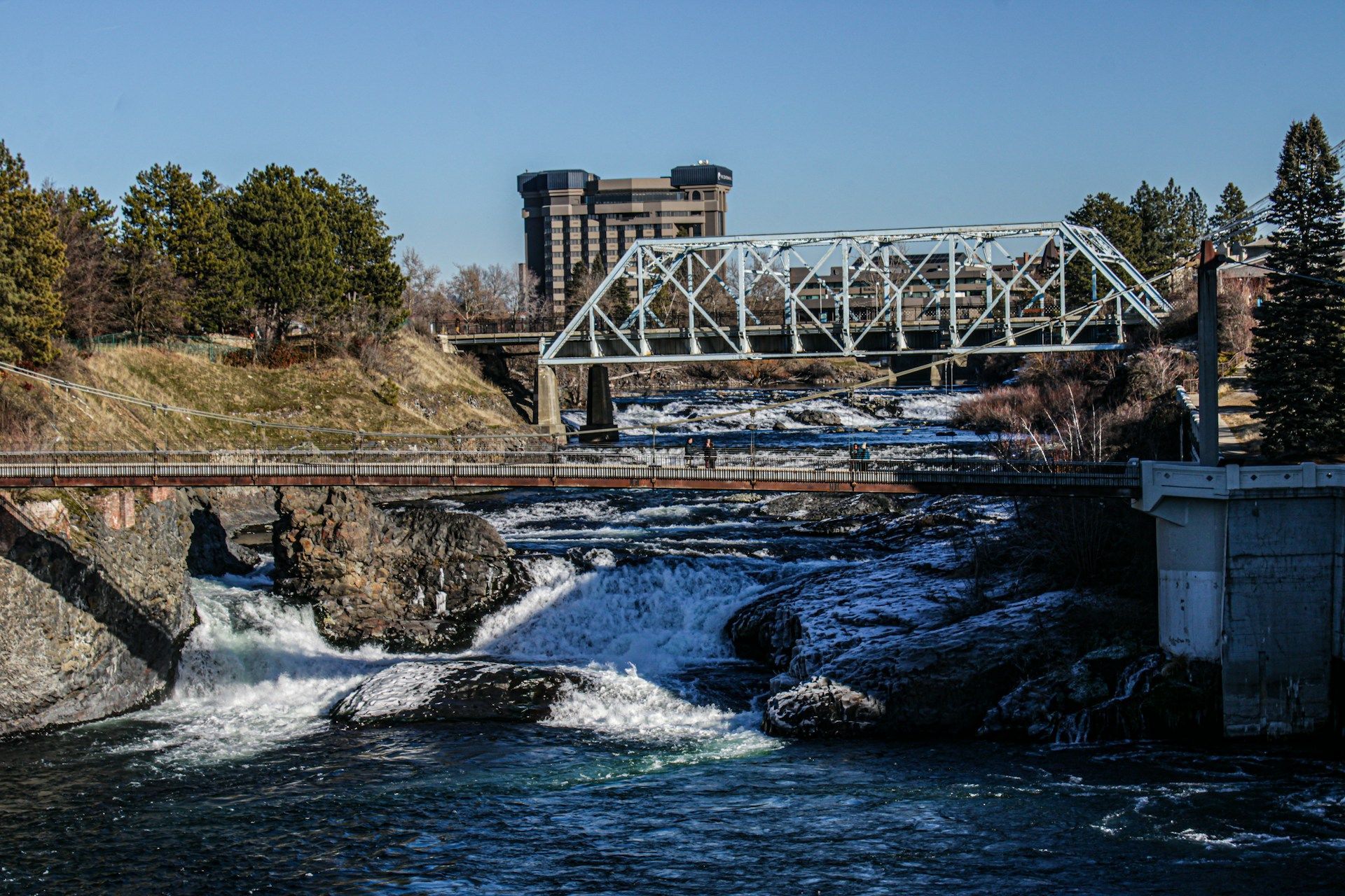A river flows under a bridge, with a castle-like building in the background; snowy banks.