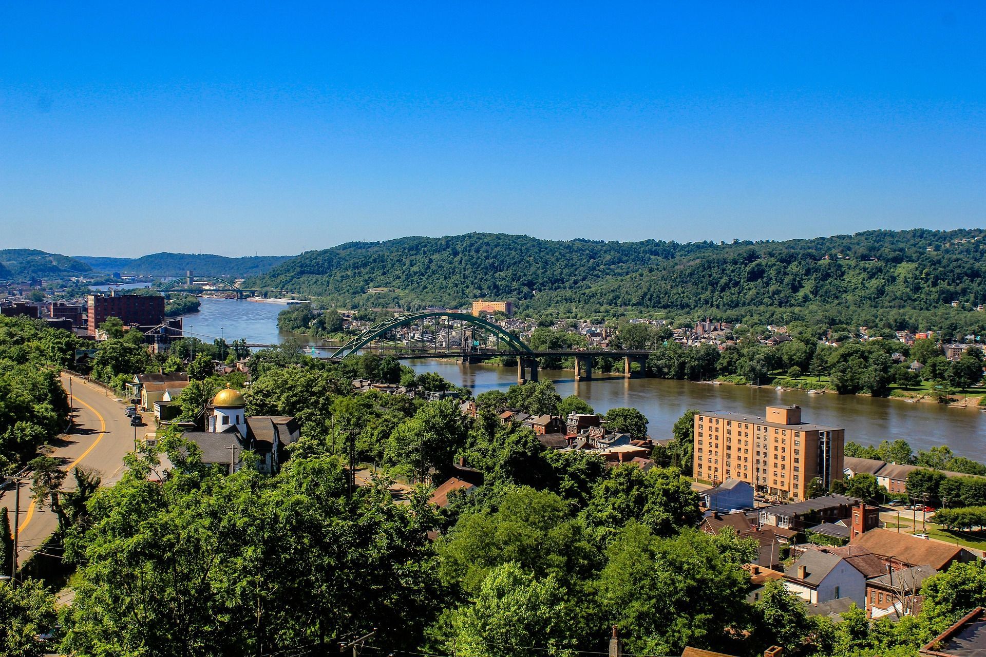 Cityscape with a river, bridge, trees, and buildings under a clear blue sky.