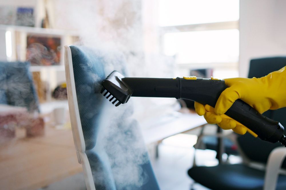 Person in yellow gloves steam cleans a blue upholstered chair in an office setting.