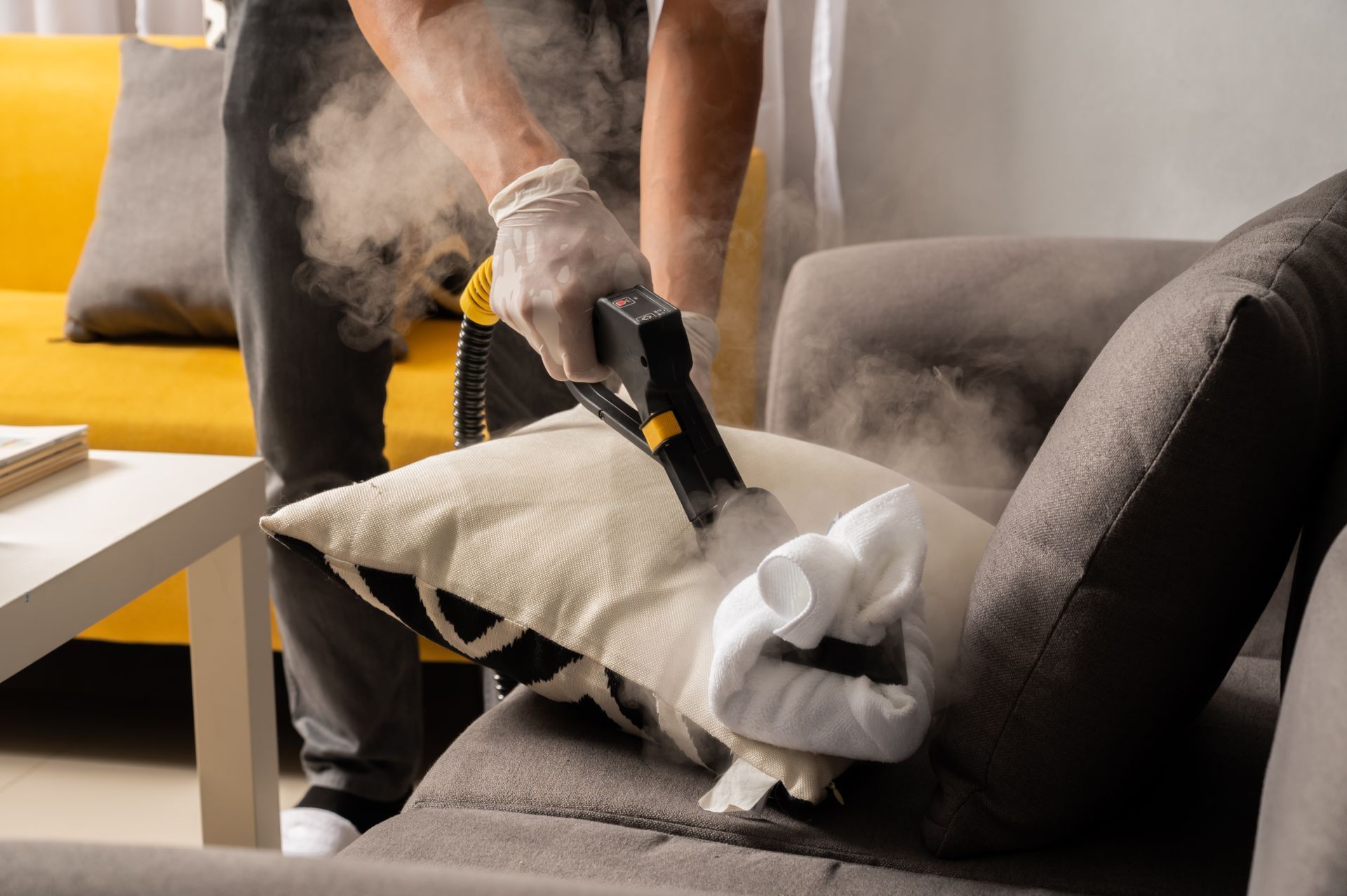 Person steam cleaning a grey couch pillow with a steam cleaner.