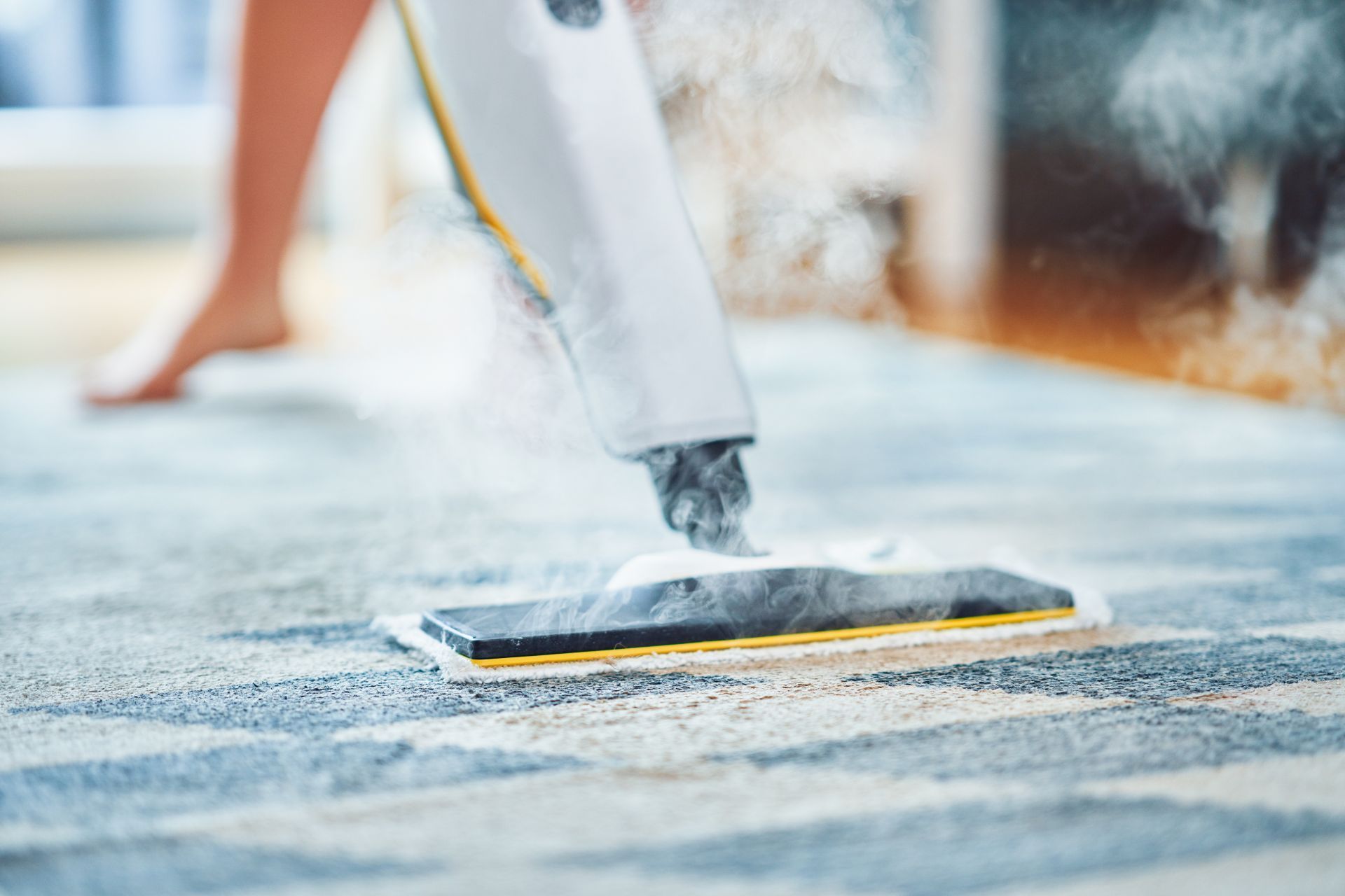 Steam cleaner being used on a patterned rug, emitting steam. Bare feet visible in background.