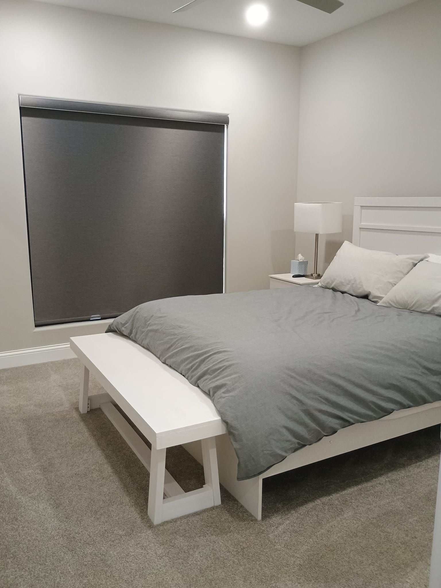 A modern bedroom with a gray bed, white nightstand, and window with a dark roller shade.