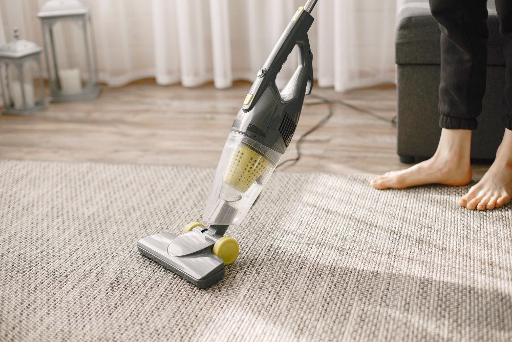 A person vacuuming a rug with a gray and yellow vacuum cleaner in a well-lit room.
